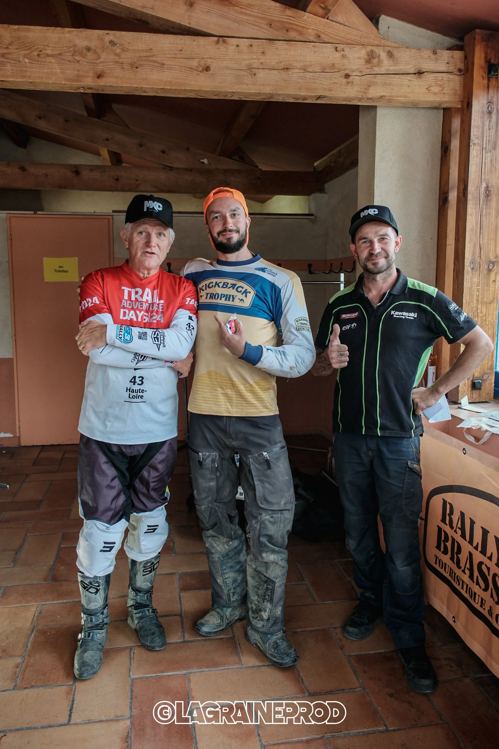 Trois hommes posent ensemble à l'intérieur, deux portant des équipements de motocross et un portant un maillot de course. Ils semblent être dans une ambiance de compétition ou d'événement sportif.