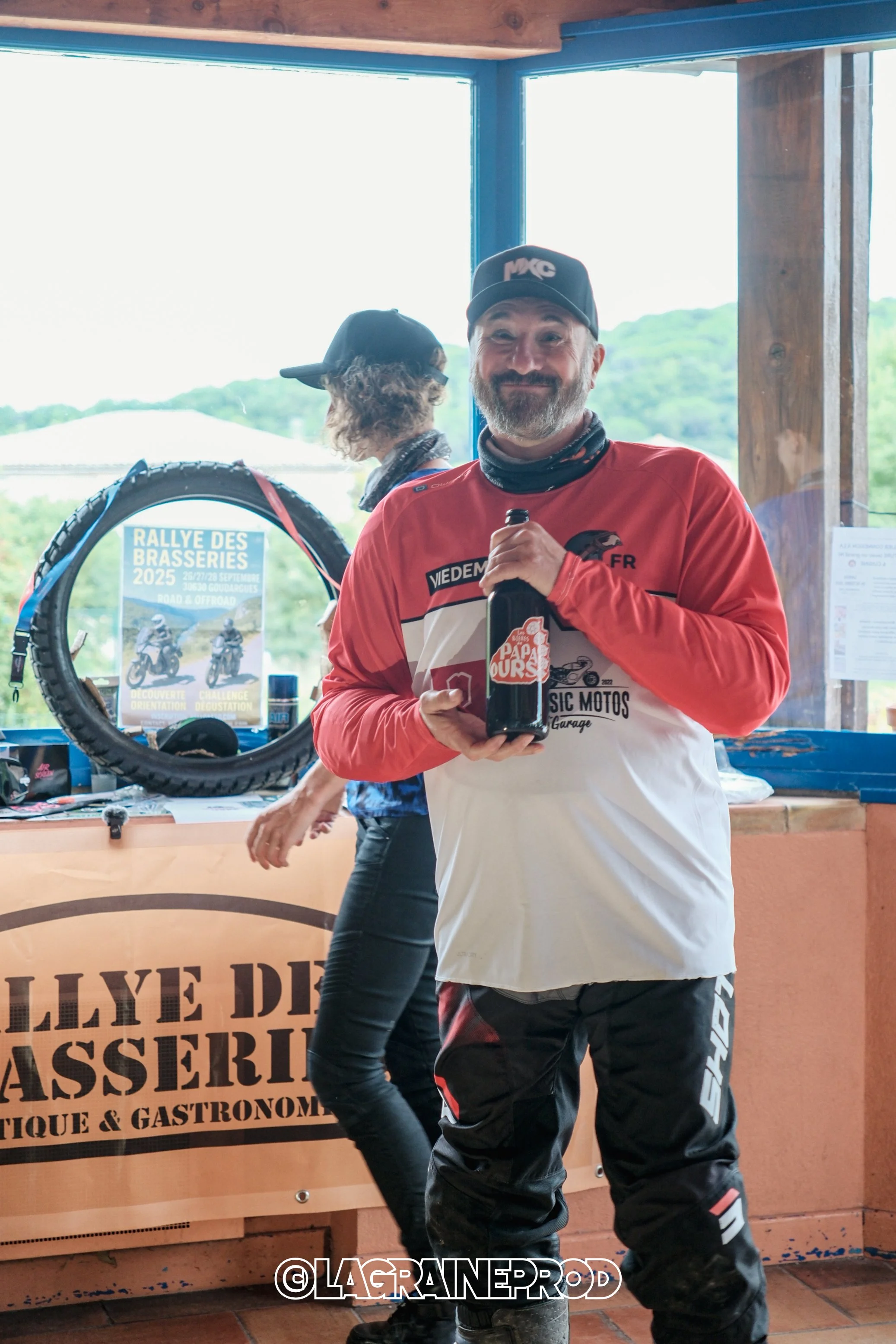 Homme souriant tenant une bouteille dans une ambiance intérieure lors d'un événement motocycliste.