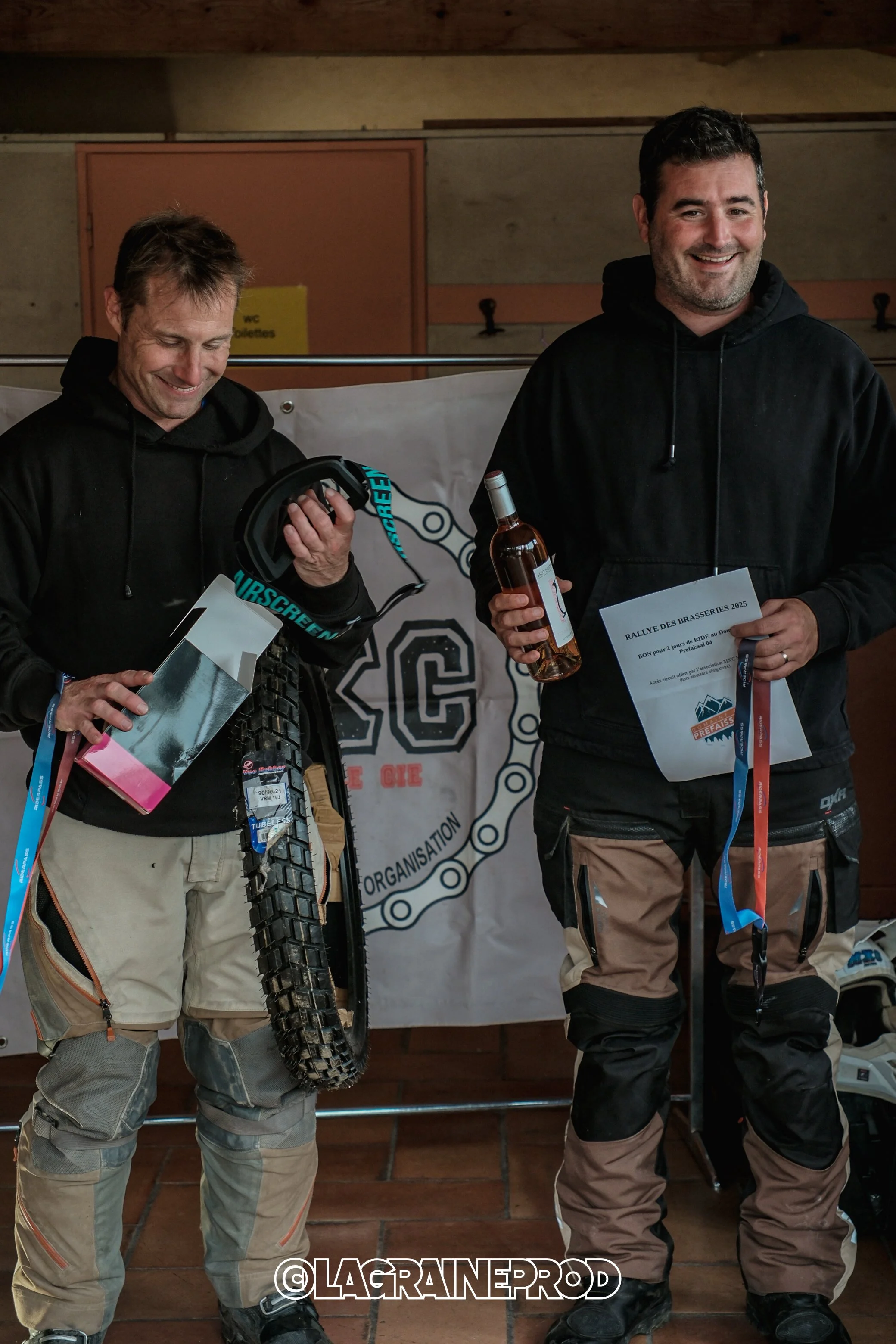 Deux hommes en tenue de moto tout-terrain, l'un tenant un casque et un pneu de moto, l'autre tenant une bouteille de boisson et un certificat, en train de sourire lors d'une remise de prix.
