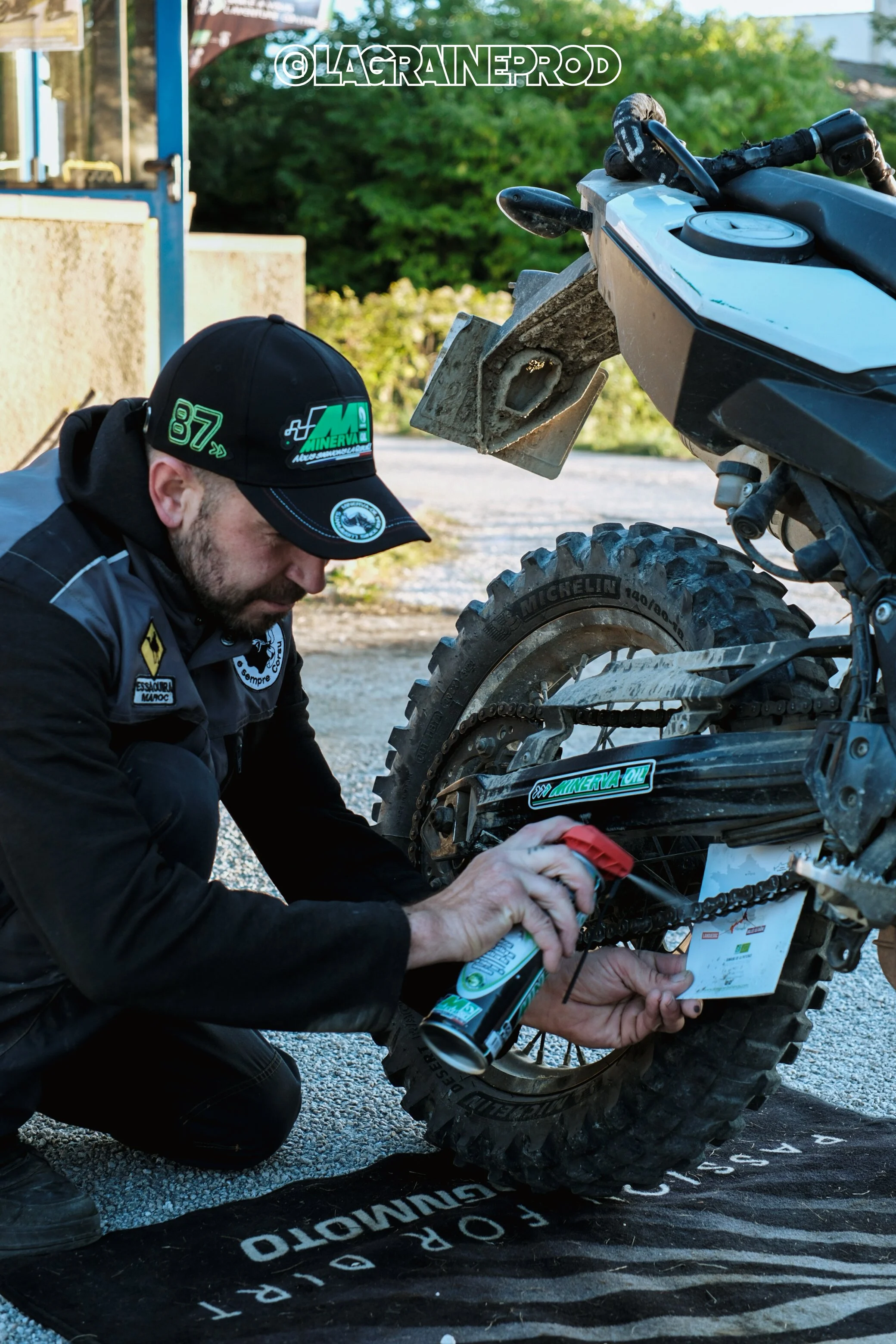 Un homme effectue une maintenance sur une moto tout-terrain, appliquant de l'huile sur la chaîne, avec un fond nature et un sol en gravier.