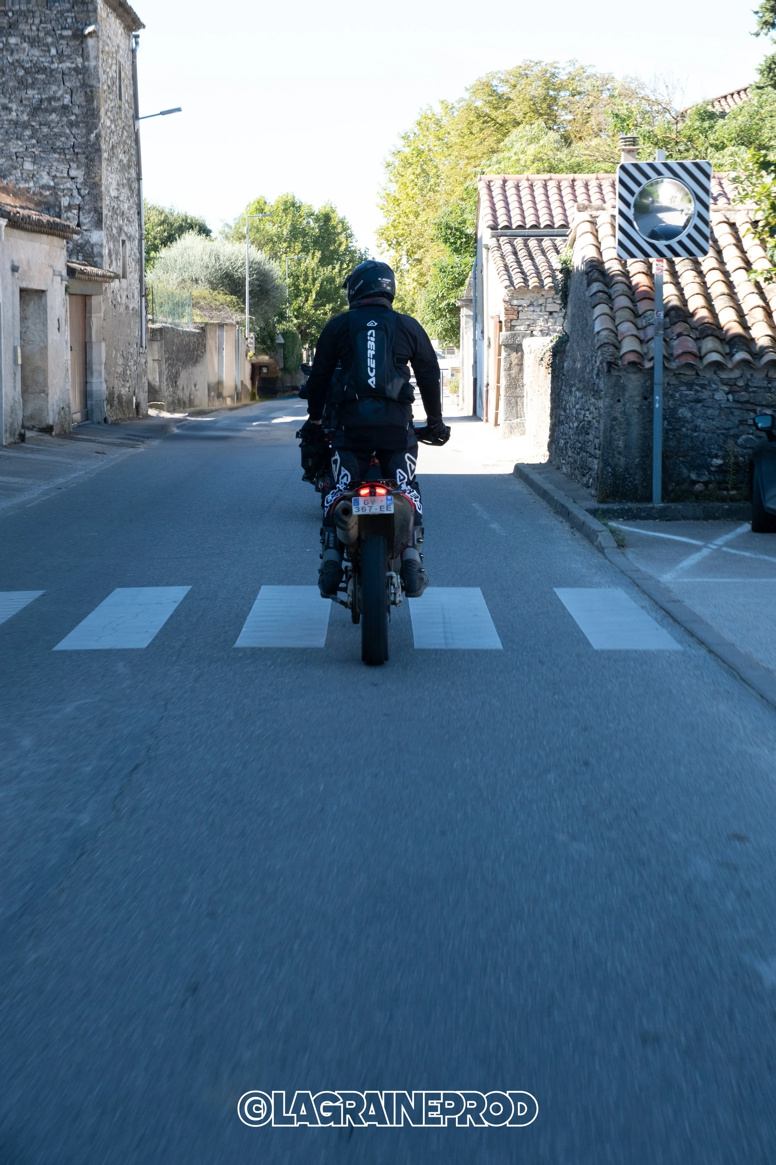 Motard en tenue noire, casque, et sac à dos, en train de rouler en moto dans une rue résidentielle avec des maisons en pierre, des toits en tuiles, et des arbres en arrière-plan.