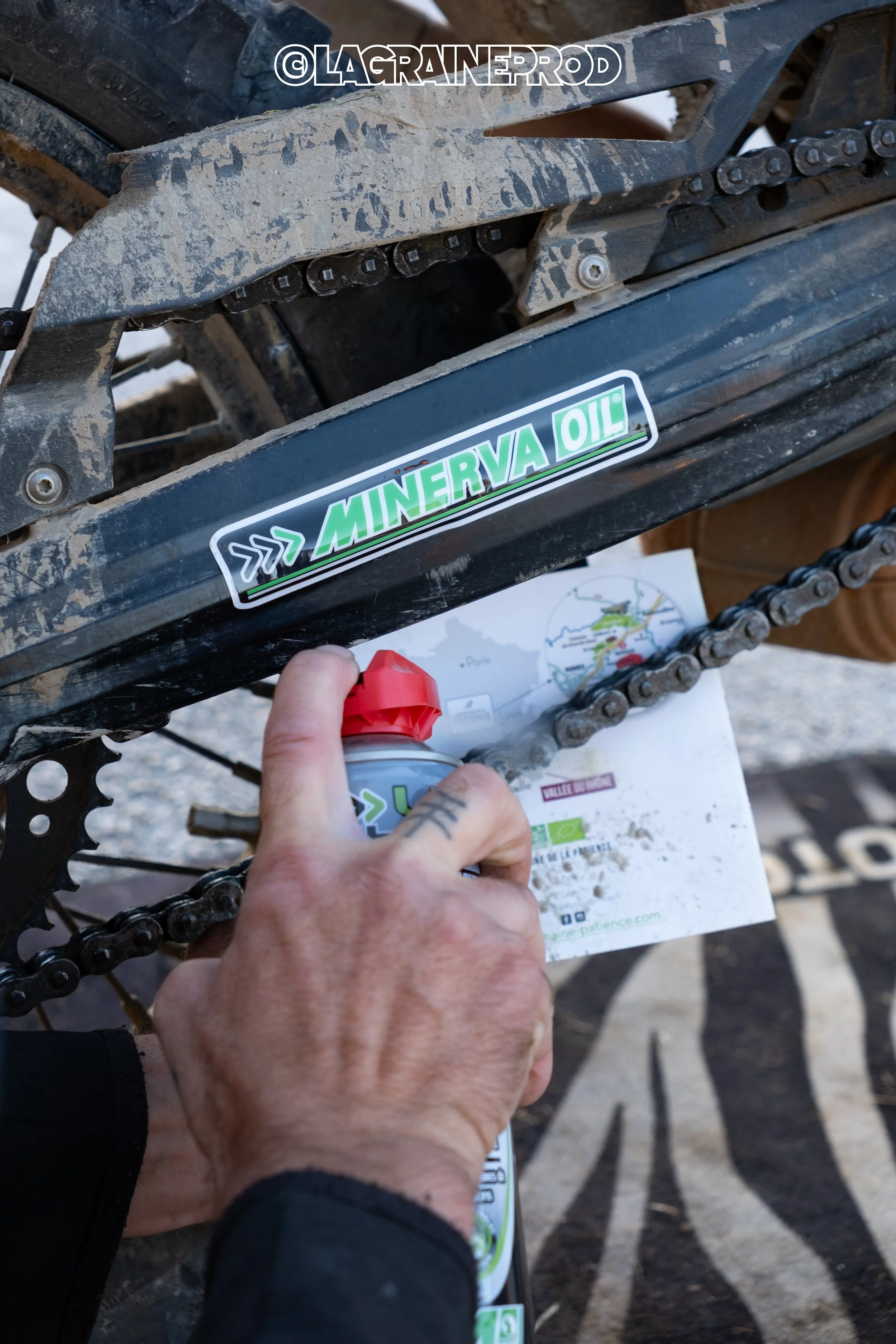 Une personne applique de l'huile Minerva Oil sur la chaîne d'une moto tout-terrain, qui est sale et en pleine action.