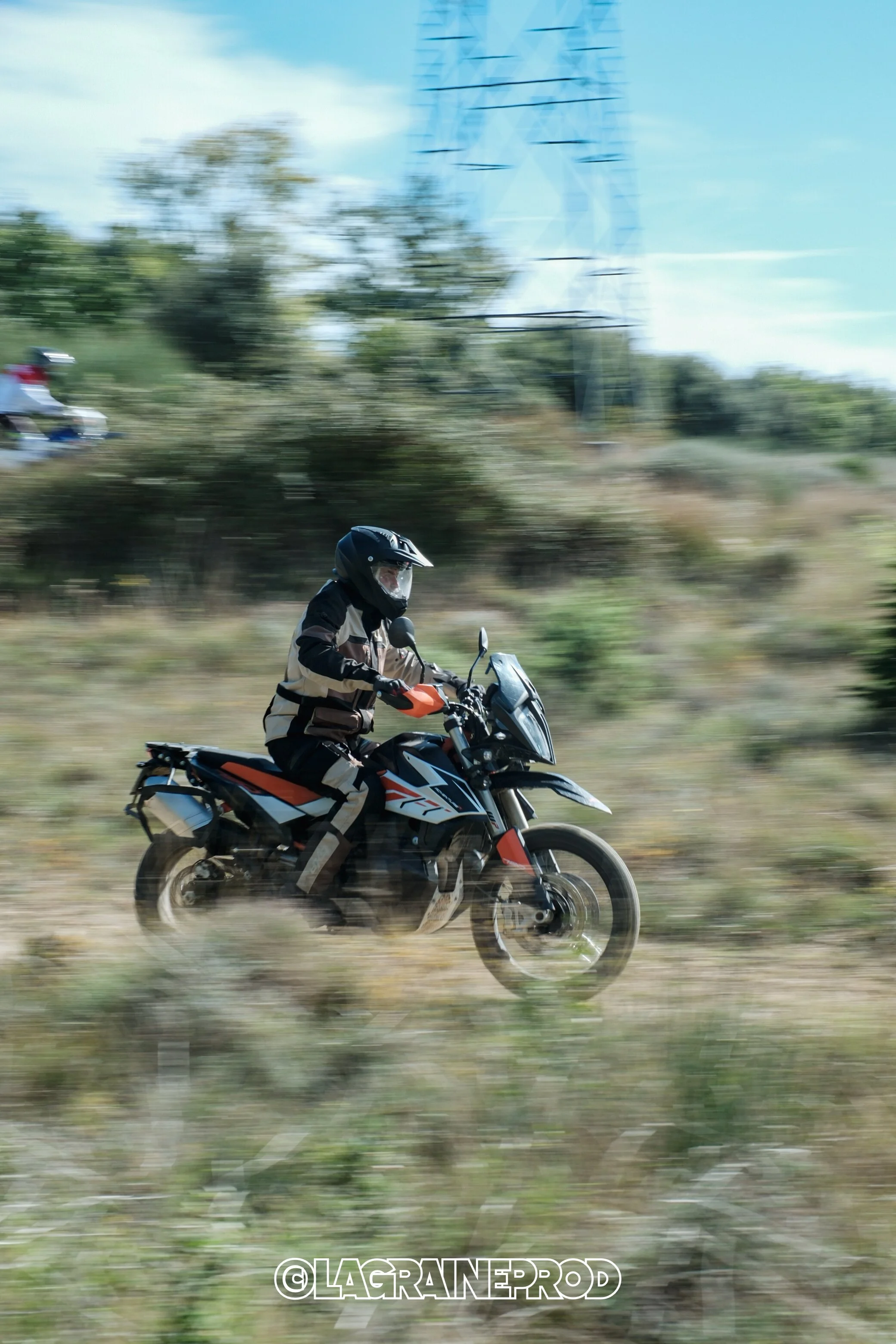 Un motard portant un casque et une veste de protection, roulant à moto dans un paysage naturel avec des herbes et des arbres, au moment où la photo a été prise en déplacement.