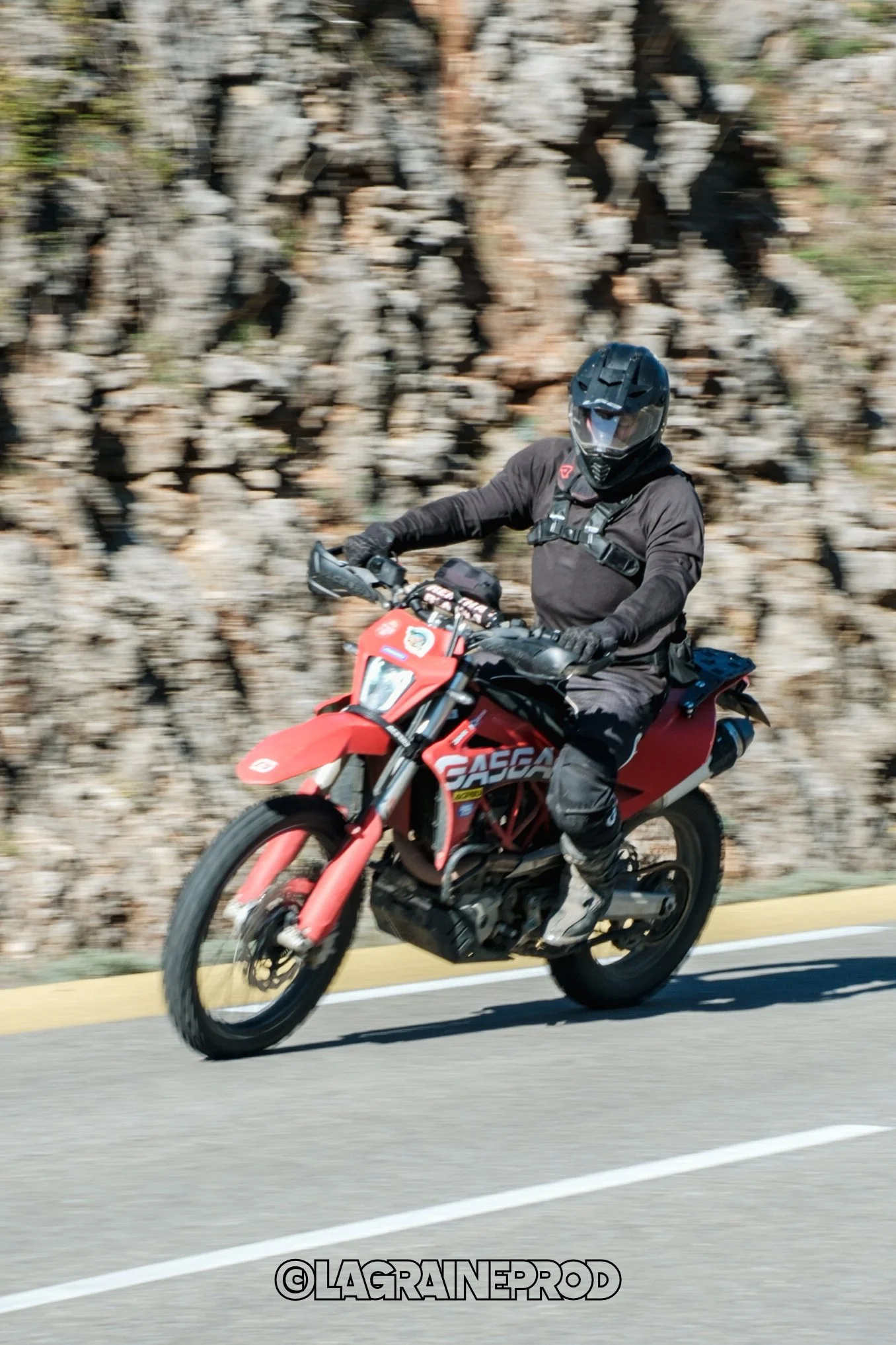 Personne portant un casque et vêtue de vêtements de moto, chevauchant une moto rouge en mouvement sur une route près d'une roche.