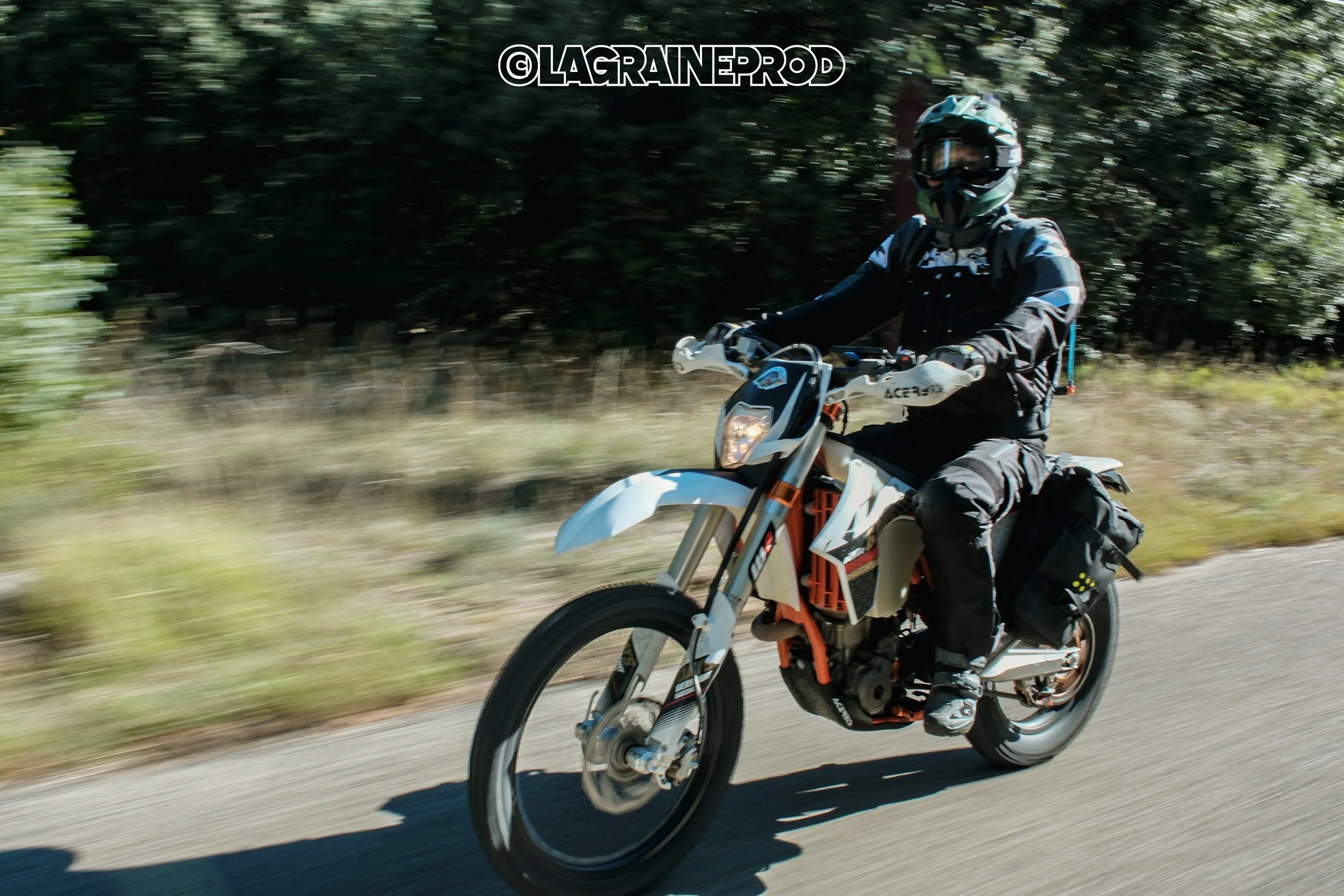 Un motard portant un casque et un équipement de protection, roulant sur une route en pleine nature, avec des arbres en arrière-plan.