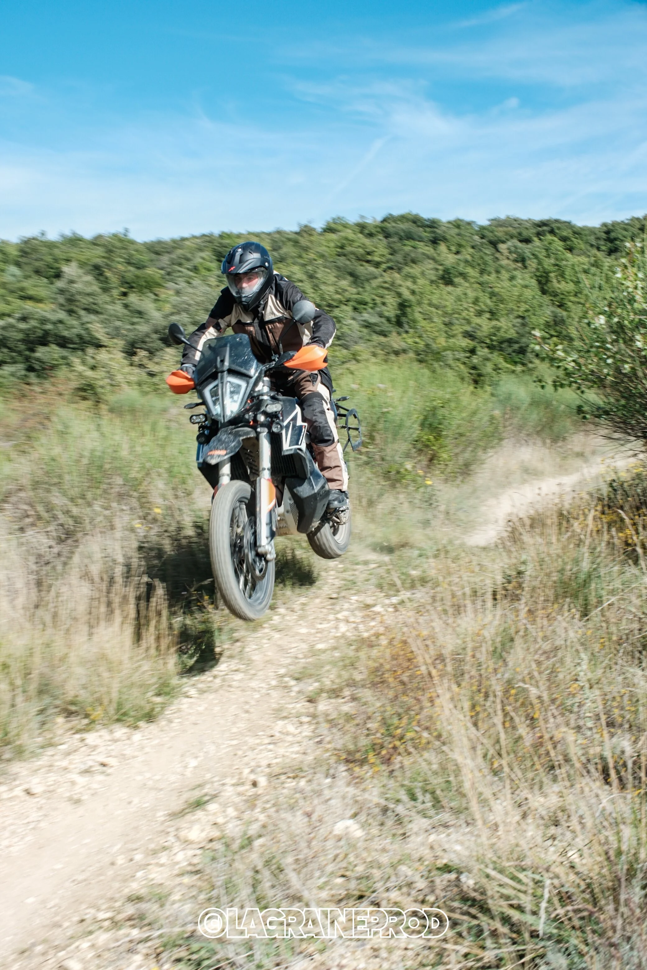 Un motard portant un casque noir et une tenue de protection, roulant à toute vitesse sur un chemin de terre à travers une zone rurale avec des arbres et un ciel bleu.