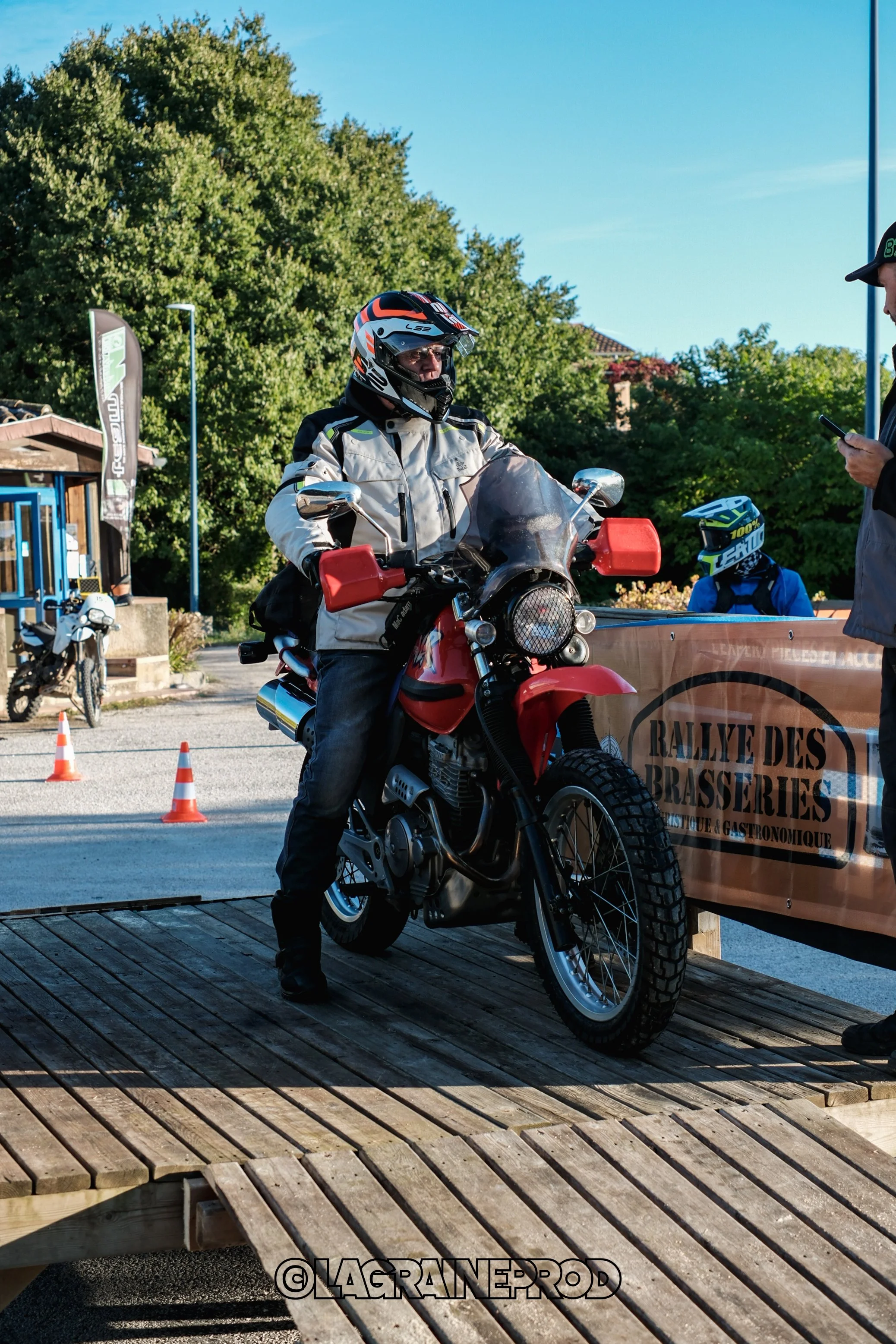 Un motard portant un casque intégral et une veste de protection monte sur une moto rouge lors d'un événement en plein air. Il y a des cônes de signalisation orange et blanc en arrière-plan, ainsi que d'autres personnes et motos.