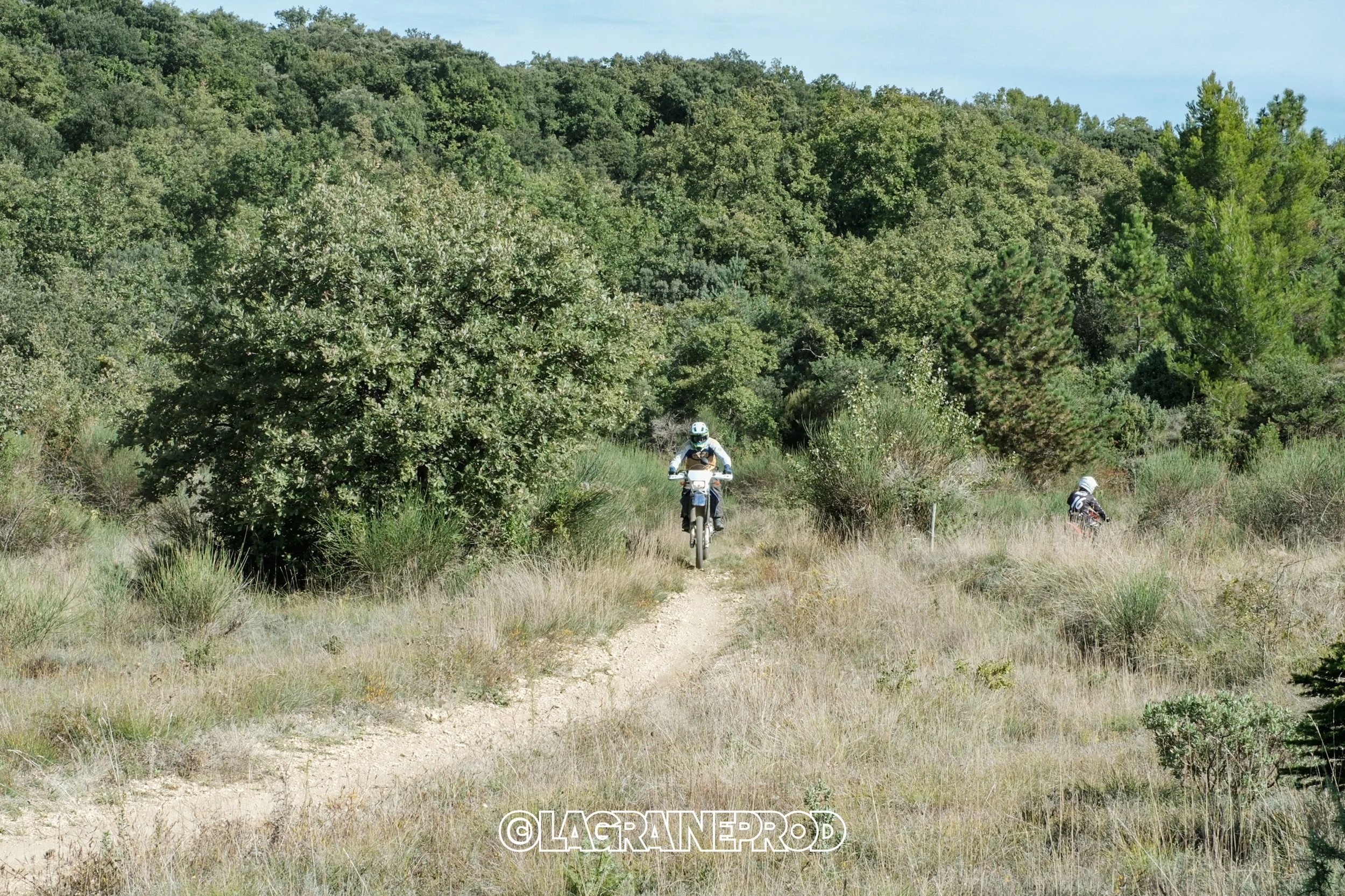 Deux motocyclistes portant des équipements de sécurité évoluent sur un sentier dans une zone boisée avec des arbres verts et un ciel clair.