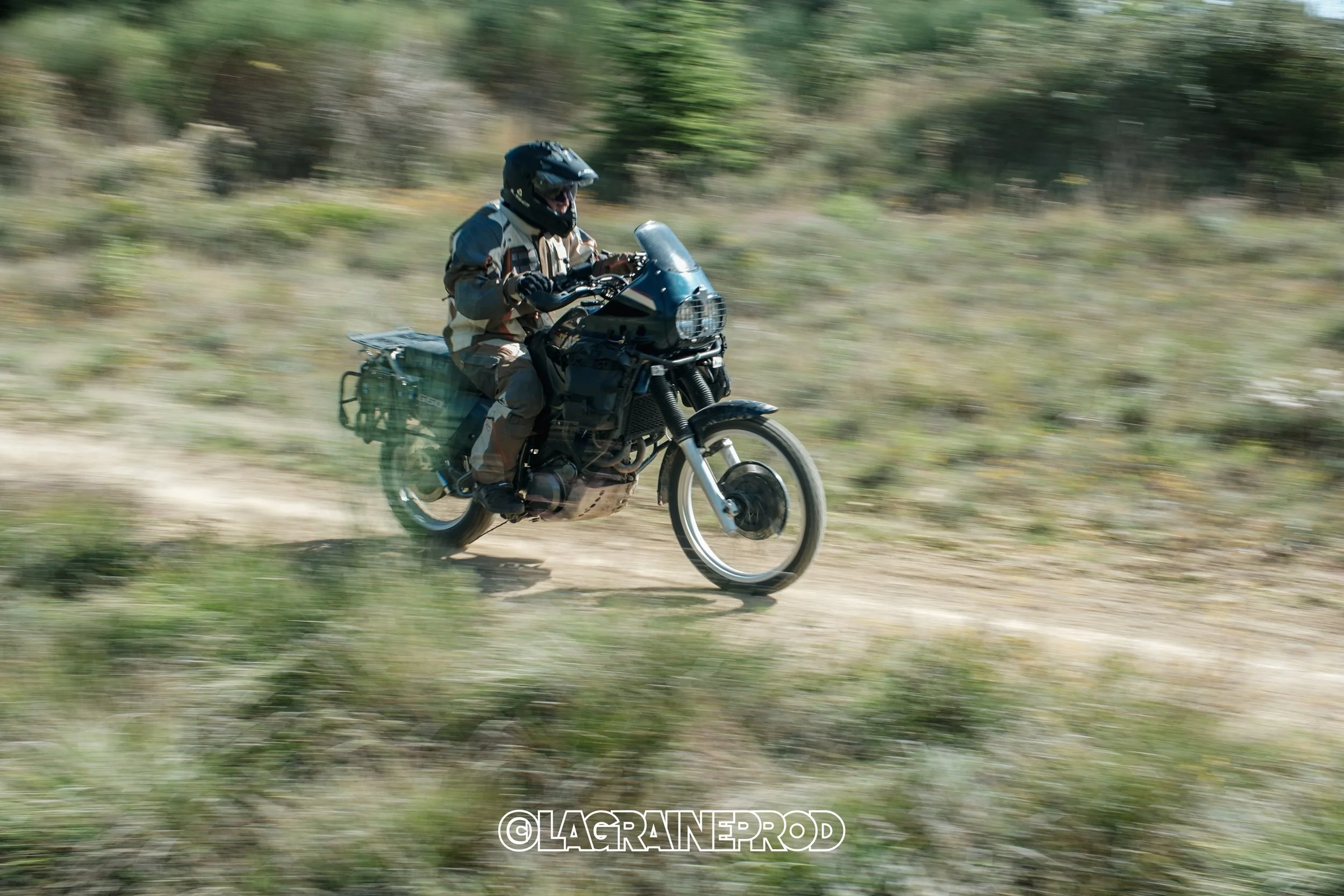 Motard portant un casque et une veste de protection, roulant rapidement sur un chemin de terre à travers un paysage boisé