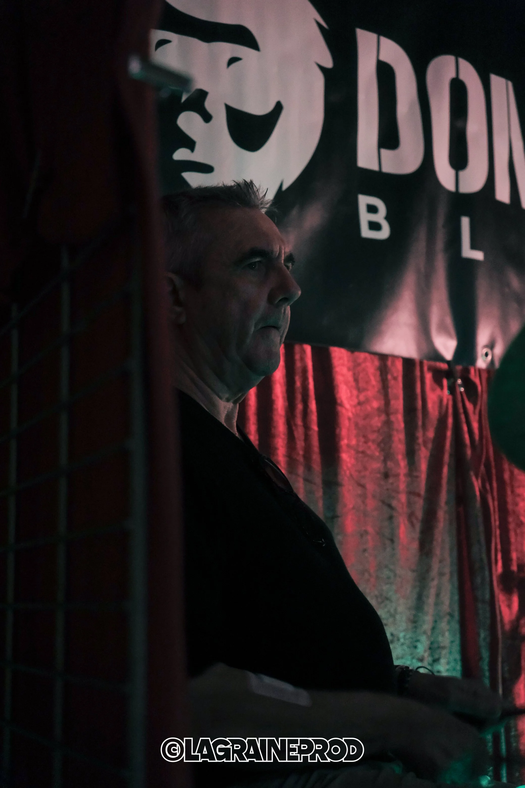 Un homme assis dans une scène sombre devant un fond avec des lettres 'DONT' et 'BL', avec un visage souriant en stylisé, en arrière-plan, et un éclairage tamisé.