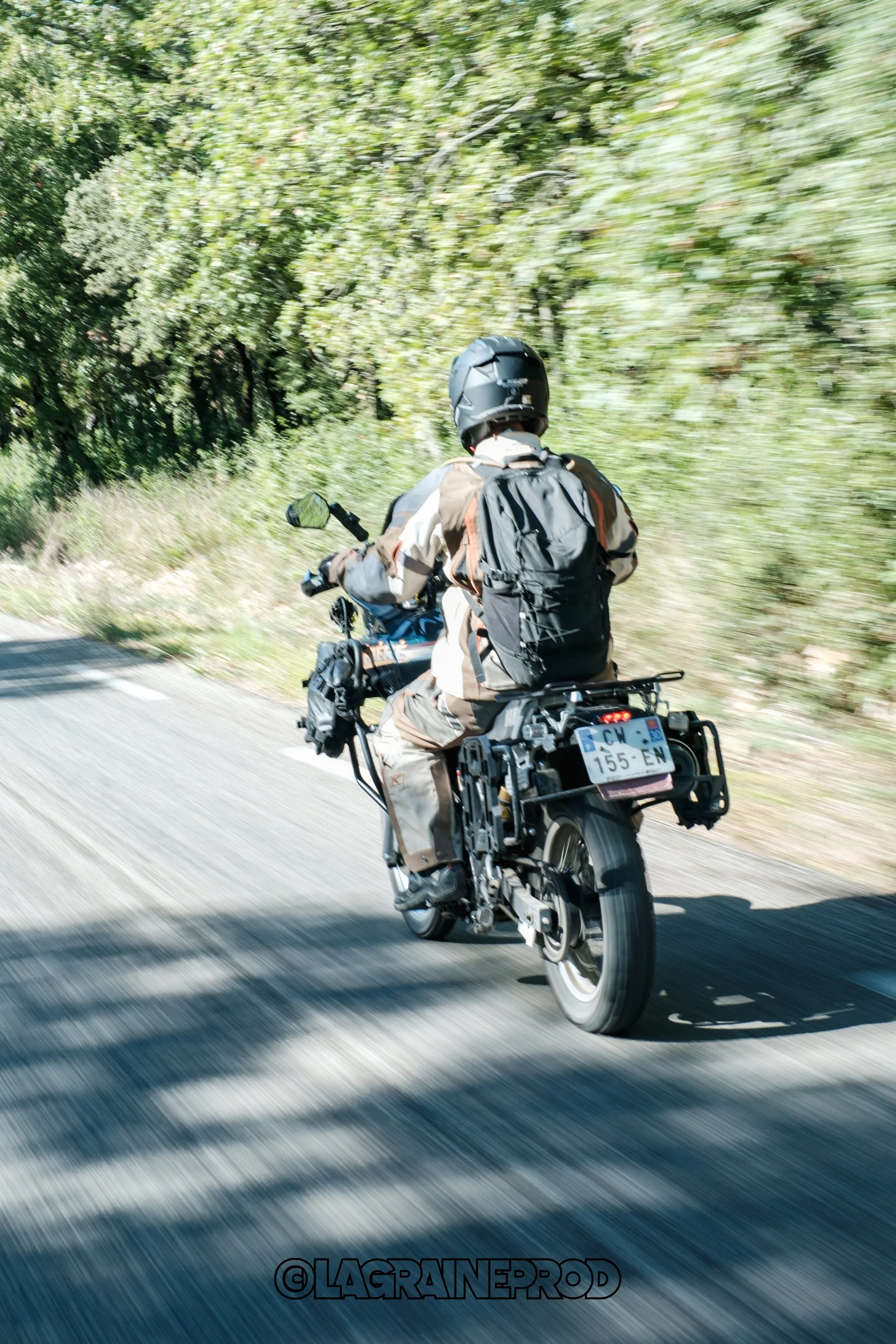 Personne en moto portant un casque et un sac à dos, voyageant sur une route entourée de végétation