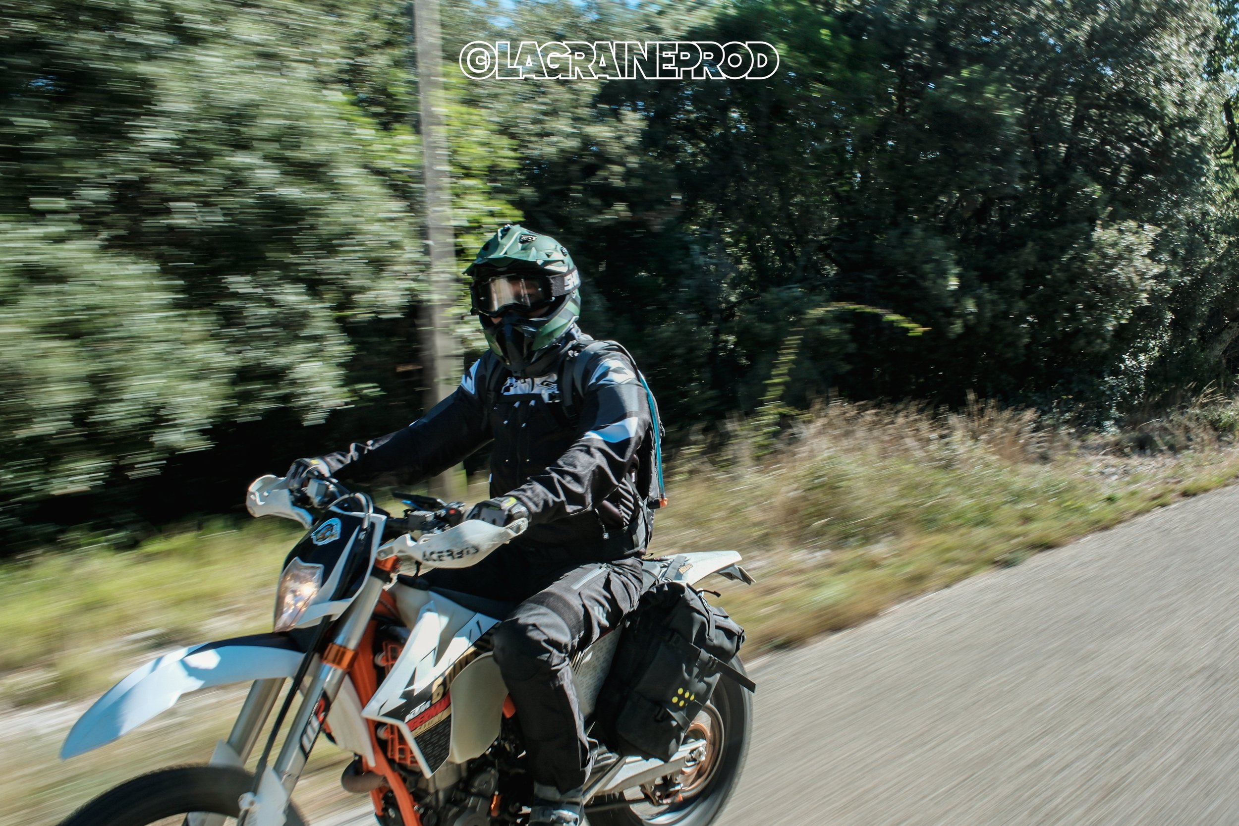 Une personne portant un casque et un équipement de moto tout-terrain, chevauchant une moto orange et blanche sur une route de campagne avec des arbres en arrière-plan.