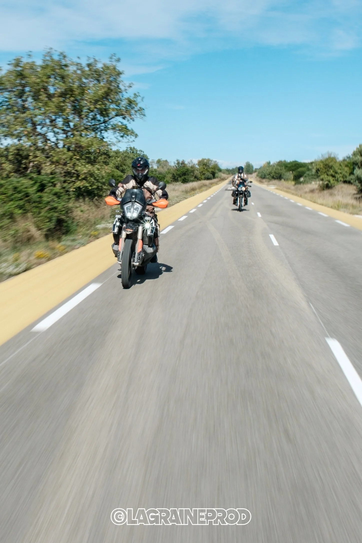 Deux motards conduisent sur une route plate, entourés de végétation sous un ciel bleu.