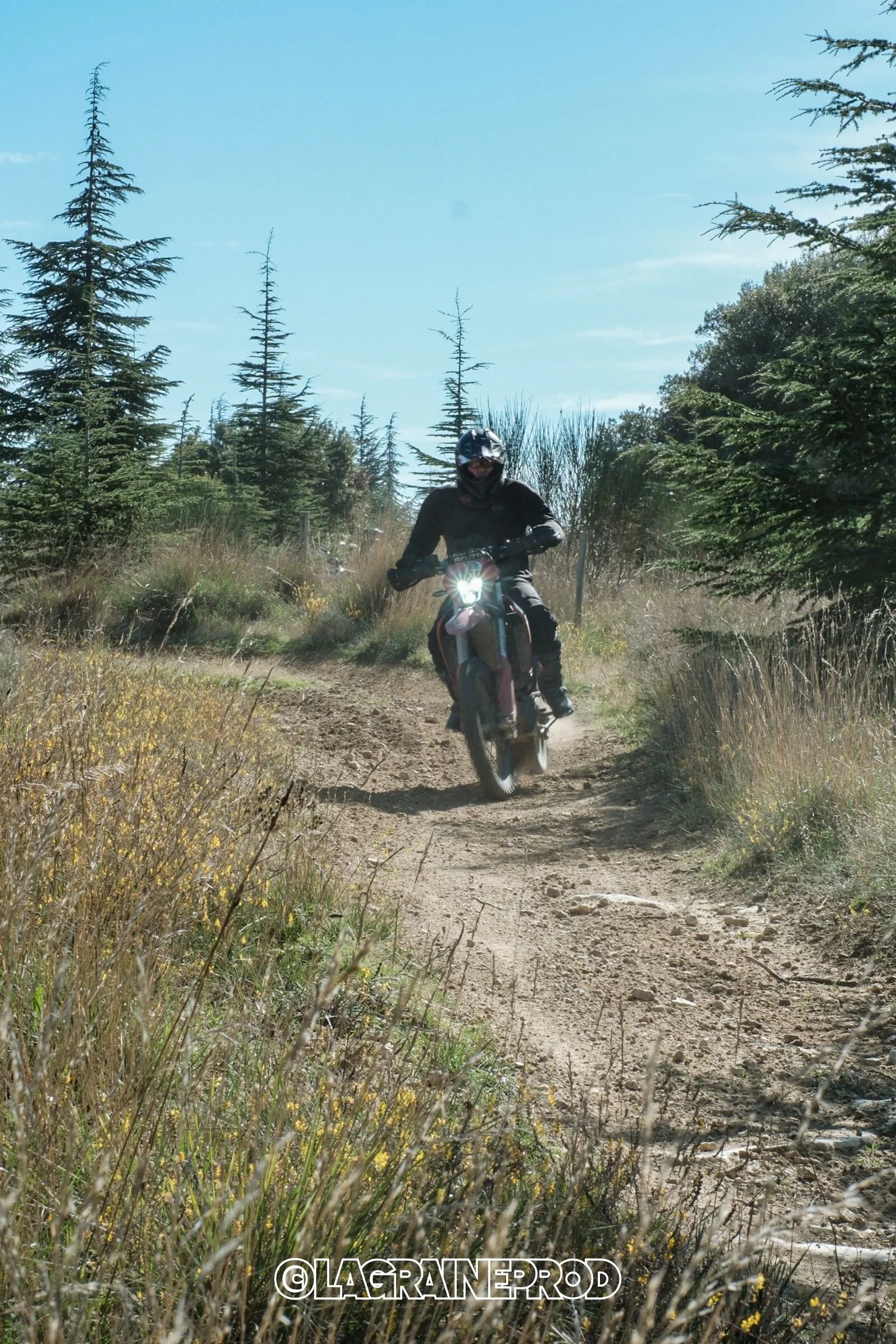 Une personne en tenue de moto tout-terrain, portant un casque, roulant sur un sentier de forêt avec des arbres verts et un ciel bleu en arrière-plan.