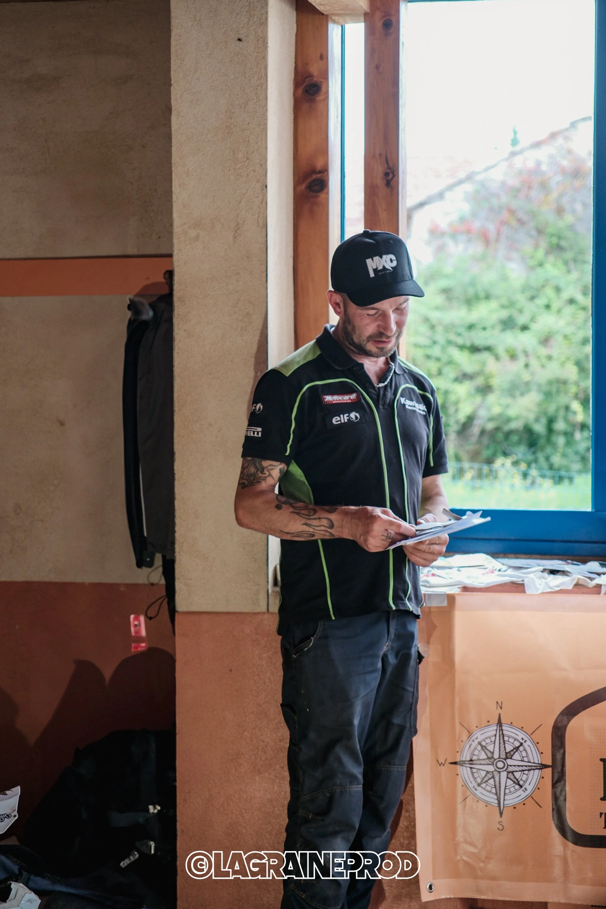 Un homme portant un t-shirt de course et une casquette, tenant un carnet, se trouve à l'intérieur près d'une fenêtre avec un arbre visible à l'extérieur.