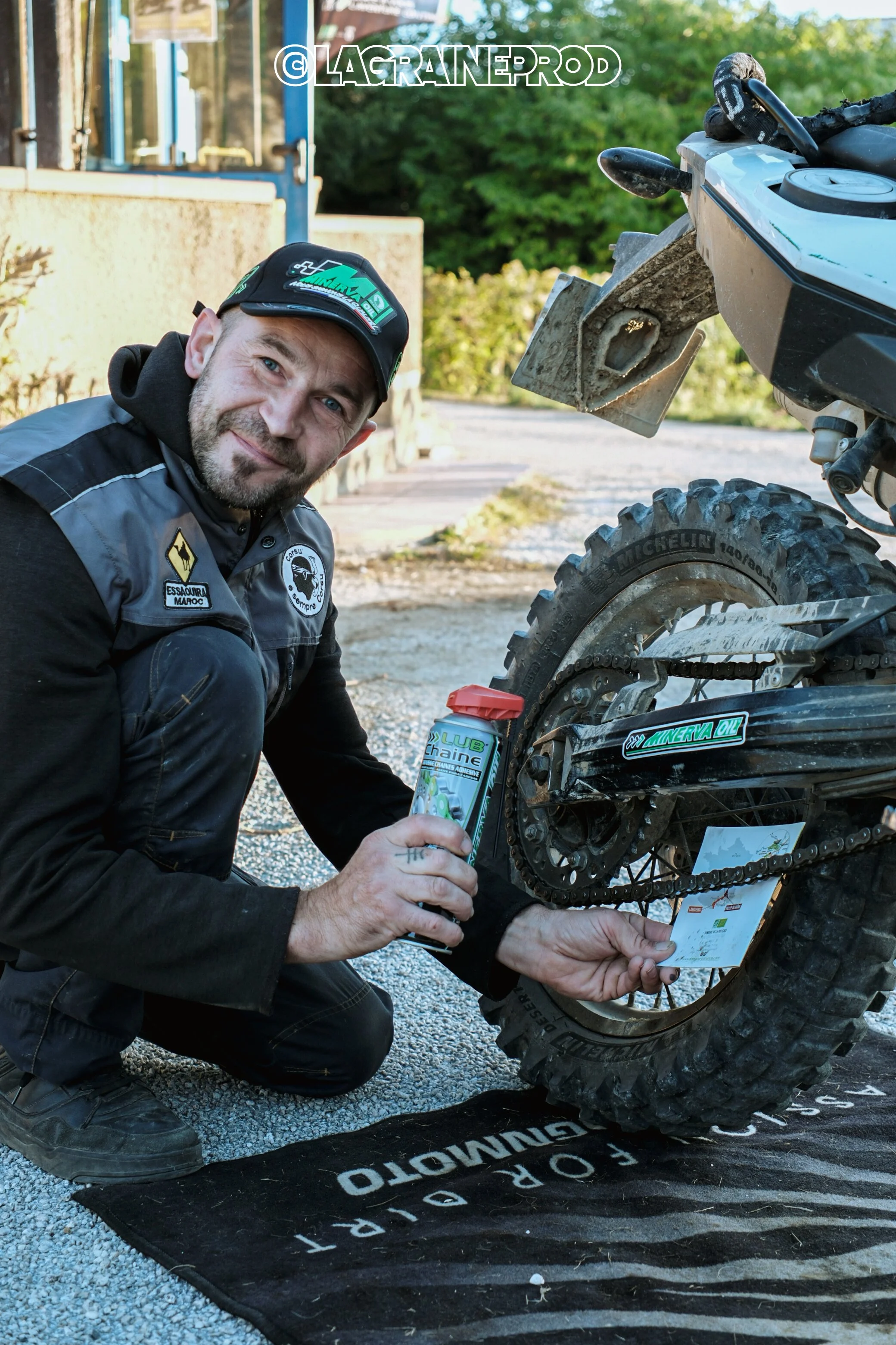 Un homme souriant en tenue de motocross, en train d'appliquer de la lubrification sur la chaîne d'une moto tout-terrain, à l'extérieur.