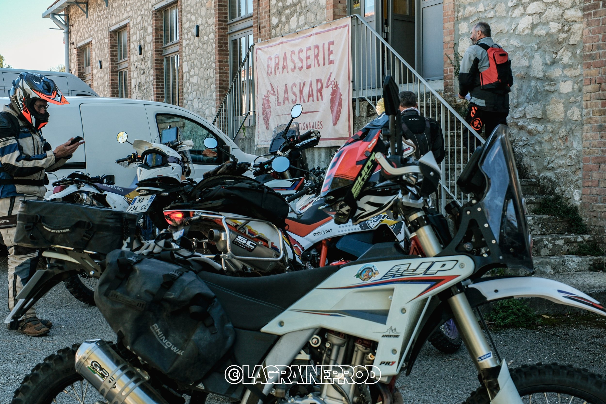 Groupe de motards avec des motos enduro et des équipements de protection, stationnés devant une brasserie artisanale appelée 'Brasserie Laskar'.