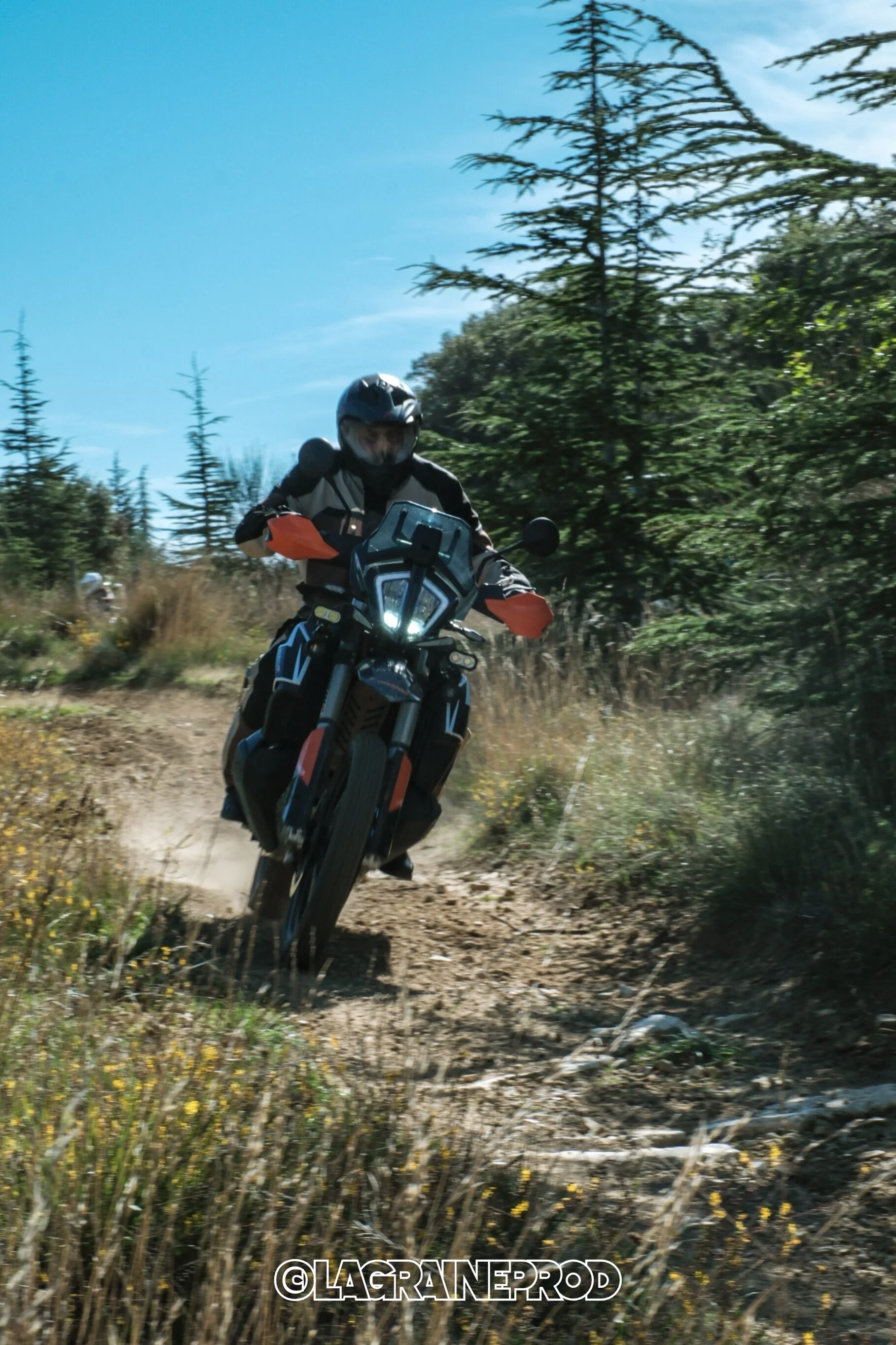 Un motard portant un casque, une veste de protection et des gants, roule sur une moto tout-terrain sur un sentier en pleine nature avec des arbres et un ciel bleu en arrière-plan.