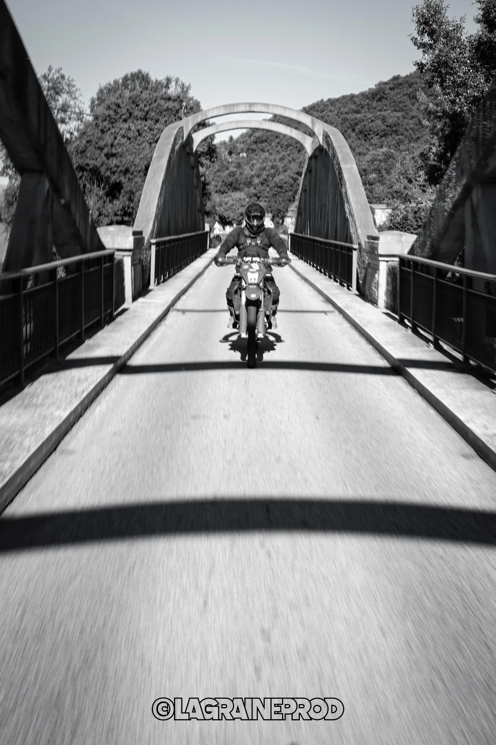 Une personne en casque et en veste pilote une moto sur un pont en arc en pierre, en noir et blanc.