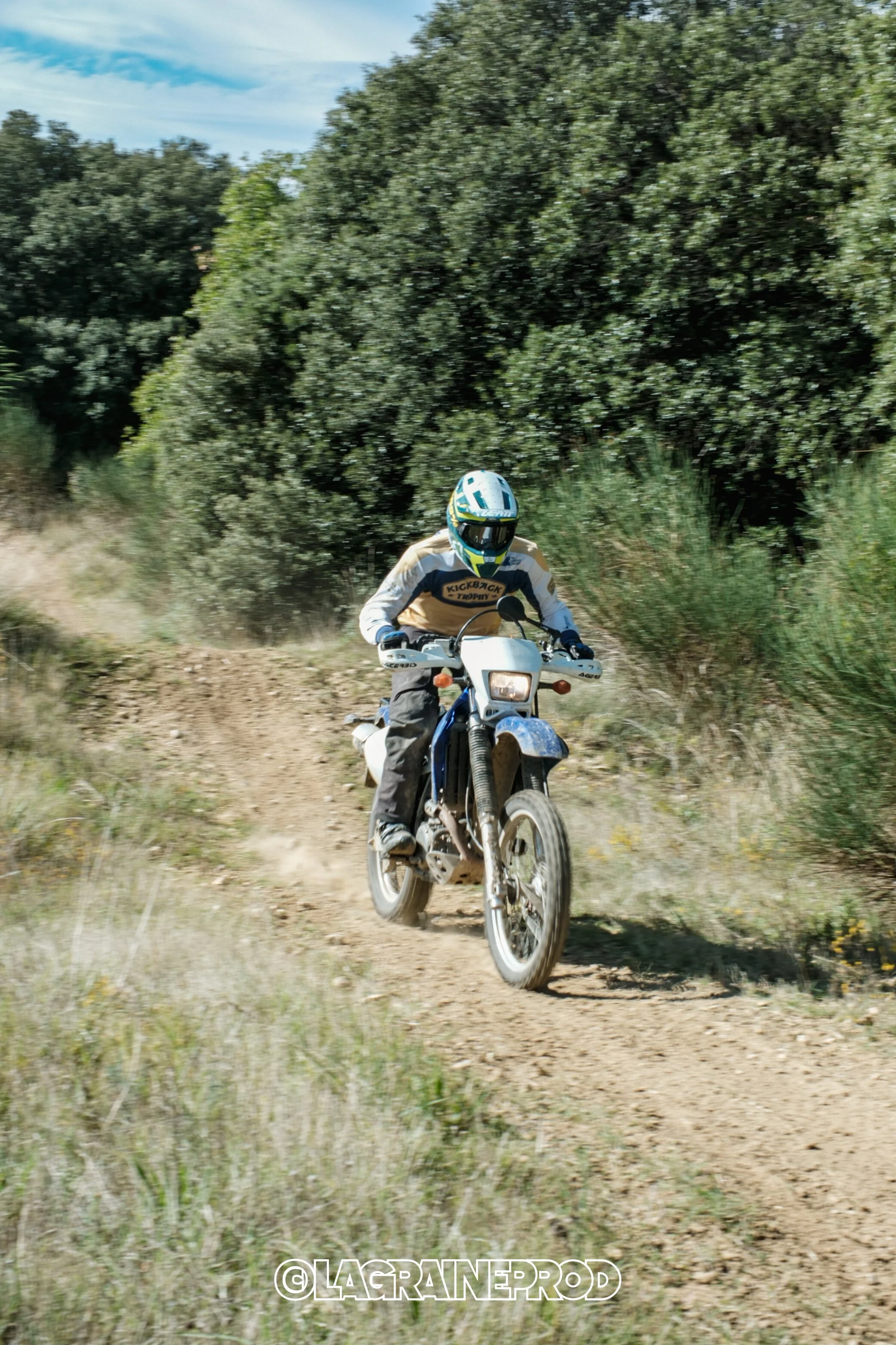 Un motocycliste en casque et vêtements de protection roule sur un sentier de terre dans un environnement herbeux et boisé.