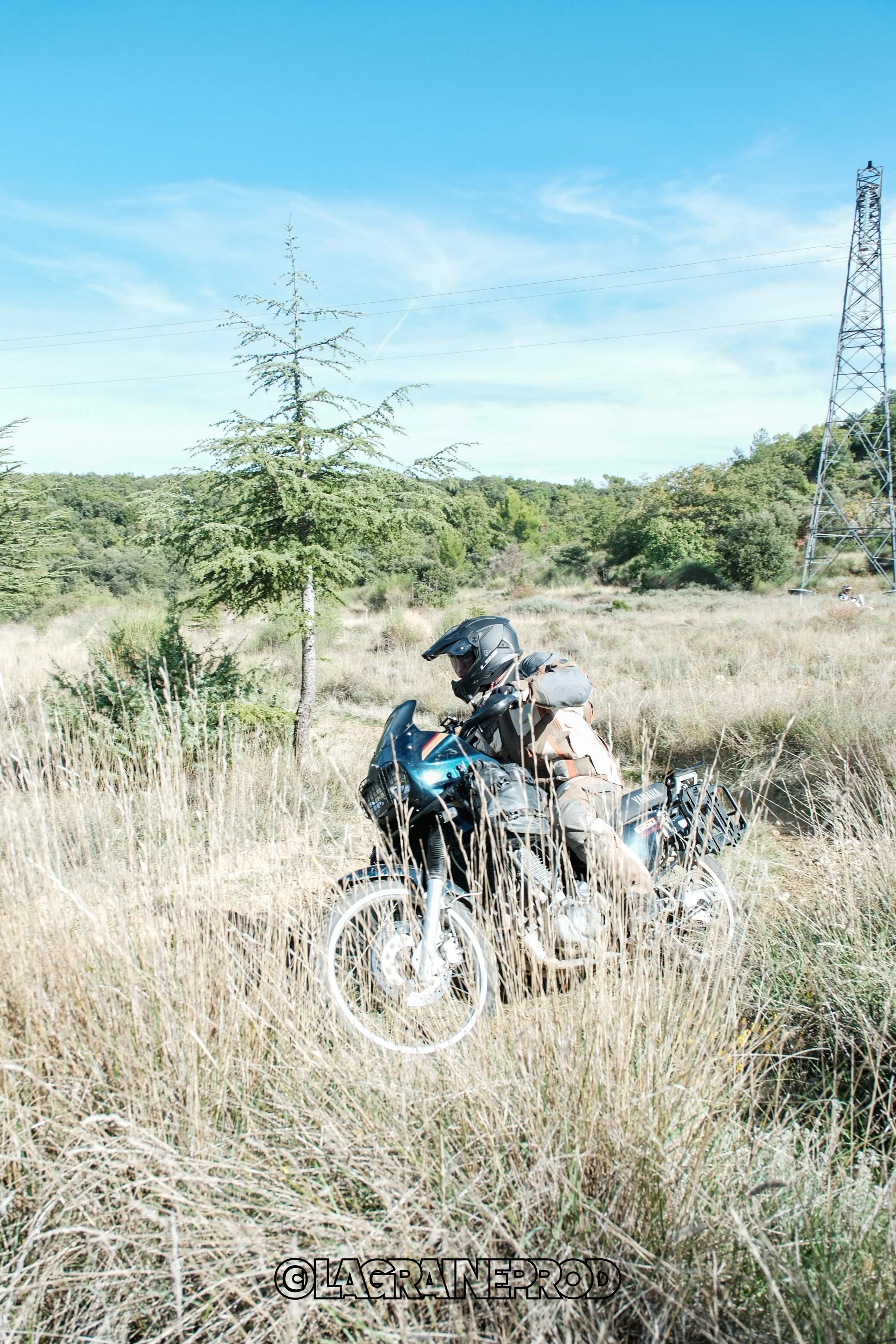 Un motard avec un casque, chargé de bagages, en train de rouler dans un champ herbeux avec des arbres et une tour électrique en arrière-plan.
