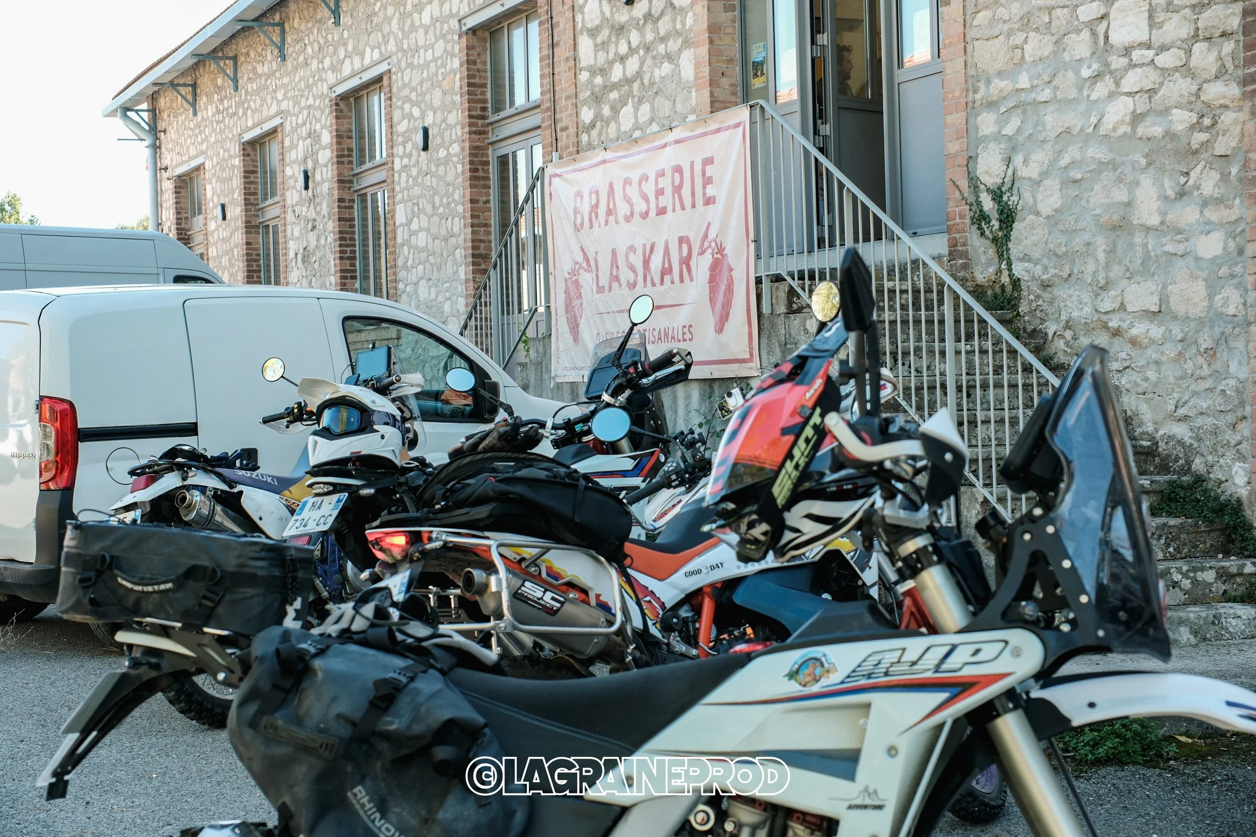 Plusieurs motos et une voiture blanche garées devant une bâtisse en pierre avec une bannière indiquant 'Brasserie Laskar'.