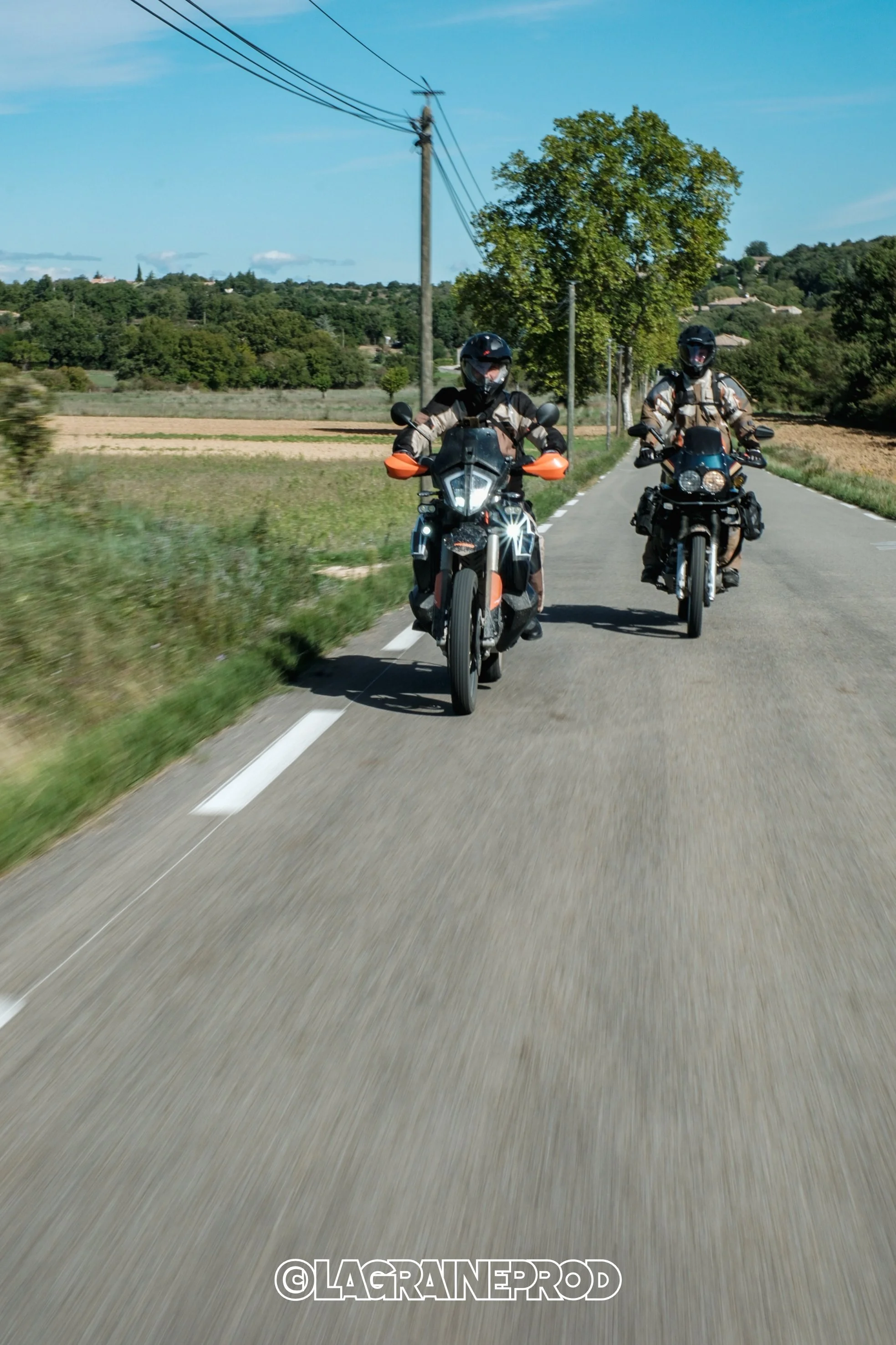 Deux motards portant des casques et des vêtements de moto roulent sur une route rurale entourée de champs et d'arbres, avec un ciel bleu.