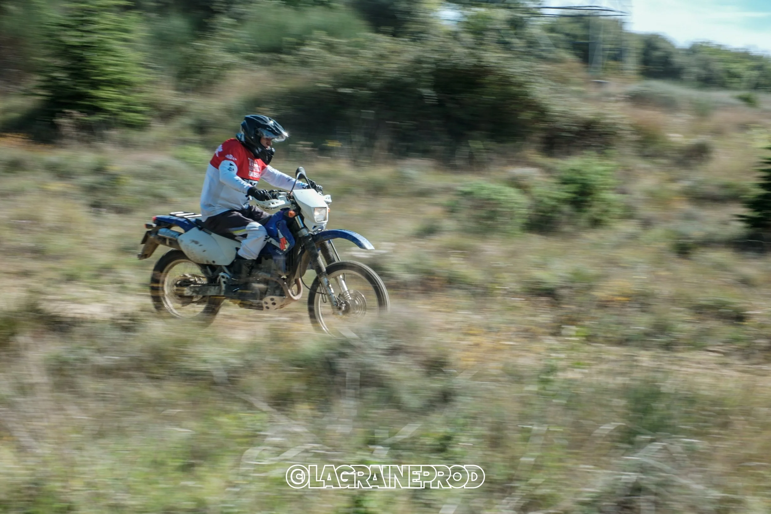 Un motocycliste portant un casque noir et une tenue blanche et rouge, roulant sur un chemin avec de la végétation verte en arrière-plan, en mouvement rapide.