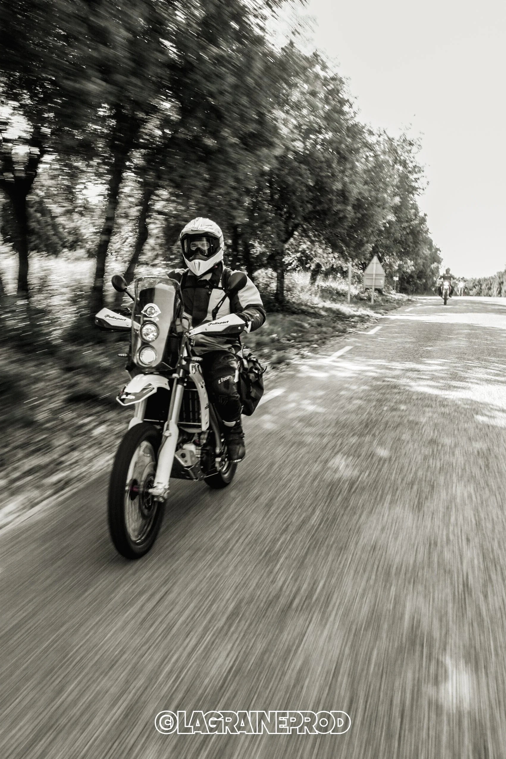 Motard portant un casque et un équipement de protection roulant à moto sur une route avec arbres en arrière-plan, image en noir et blanc.