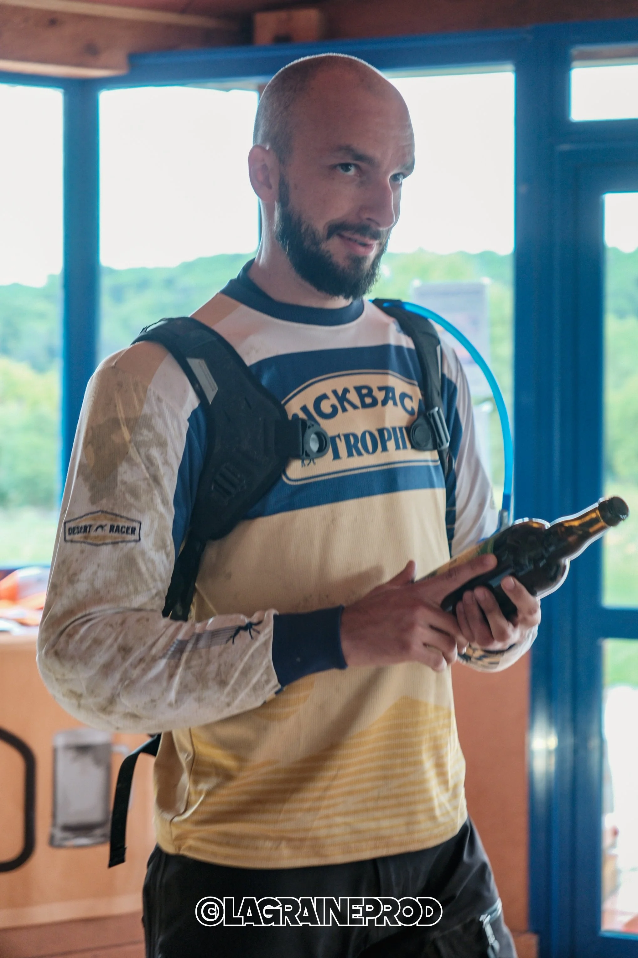 Un homme portant un maillot de motocross et un sac à dos, tenant une bouteille en verre, à l'intérieur d'un bâtiment avec de grandes fenêtres laissant voir un paysage vert.