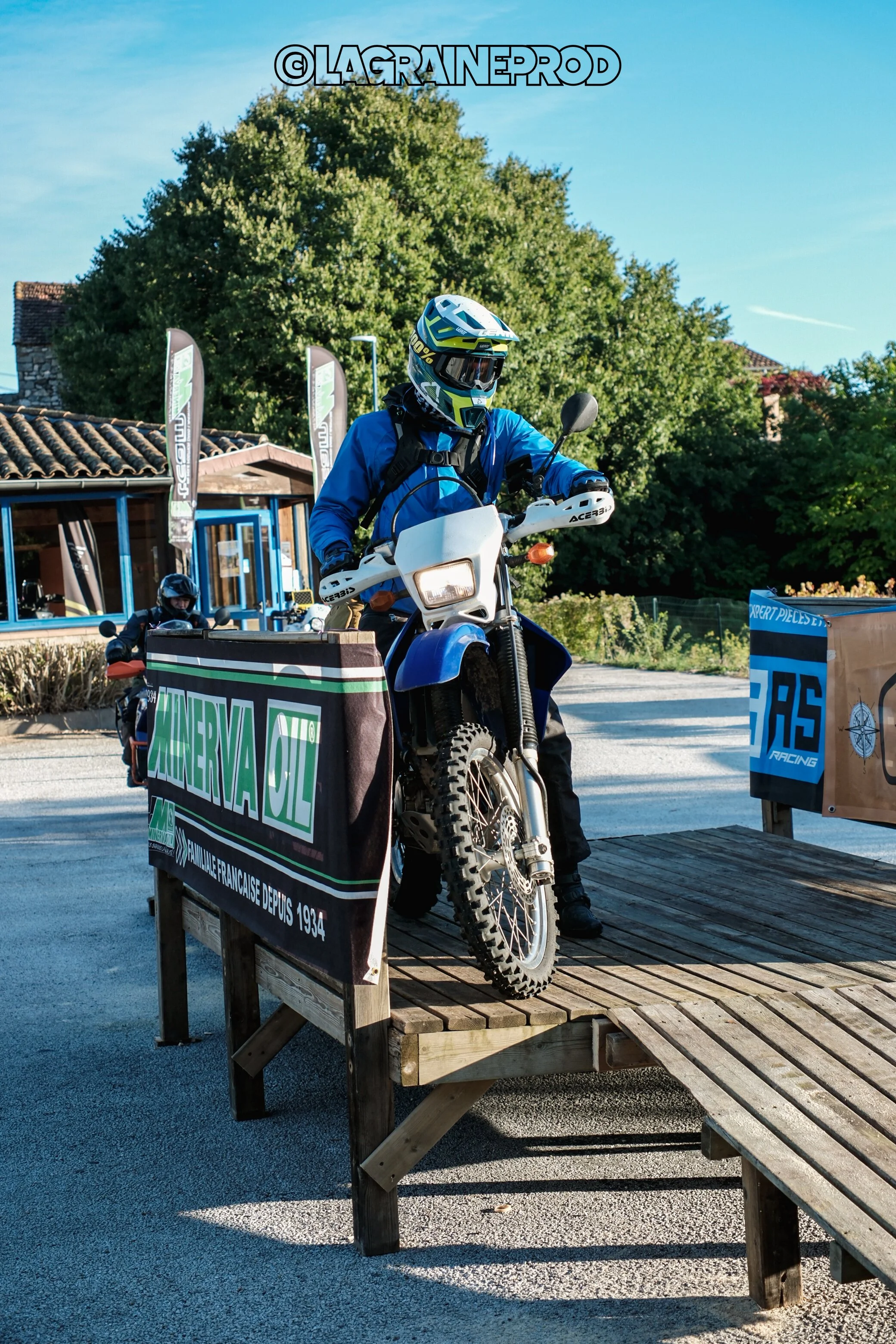 Un motard en équipement de protection, portant un casque, monte une moto tout-terrain sur une plateforme en bois lors d'un événement en extérieur, avec des panneaux et des arbres en arrière-plan.