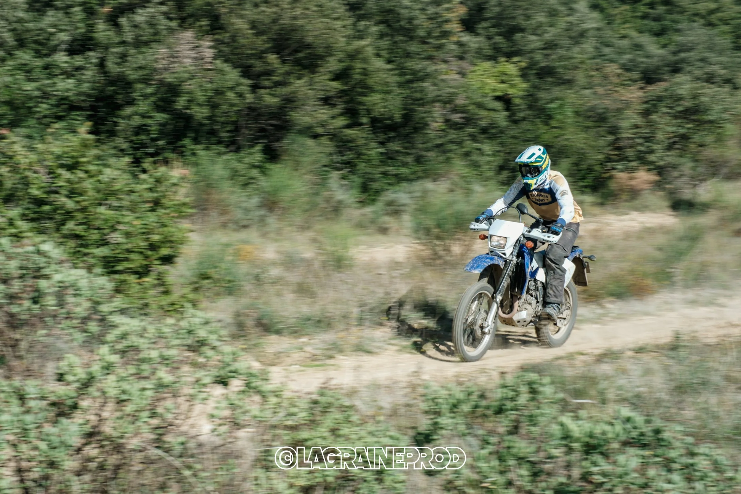Une personne en casque et équipement de protection roulant à moto tout-terrain sur un sentier poussiéreux en milieu naturel, entourée d'arbustes et d'arbres.