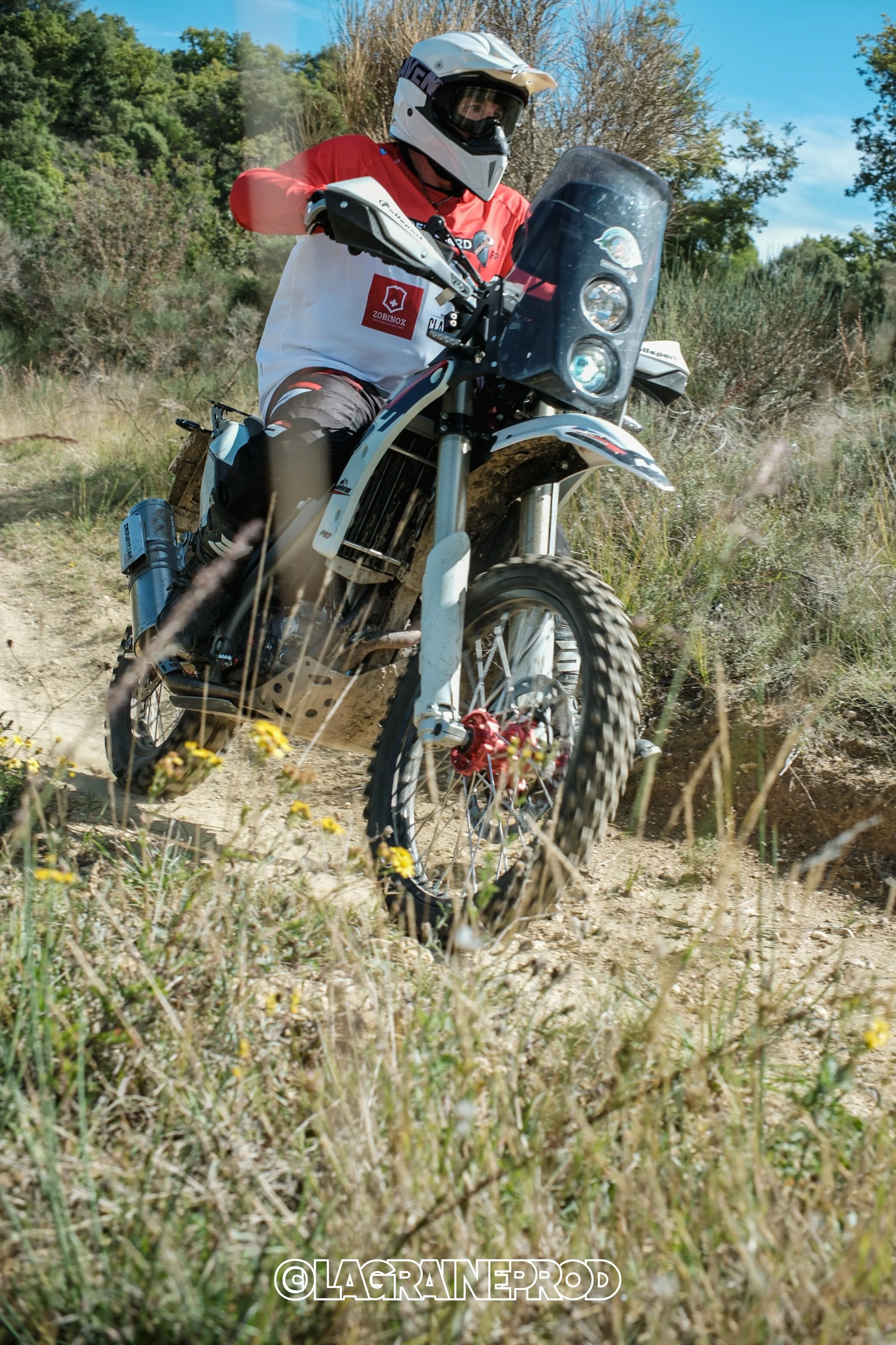 Personne faisant de la moto tout-terrain avec un casque blanc, portant un maillot blanc et rouge, sur un sentier de terre avec des herbes et des arbres derrière.