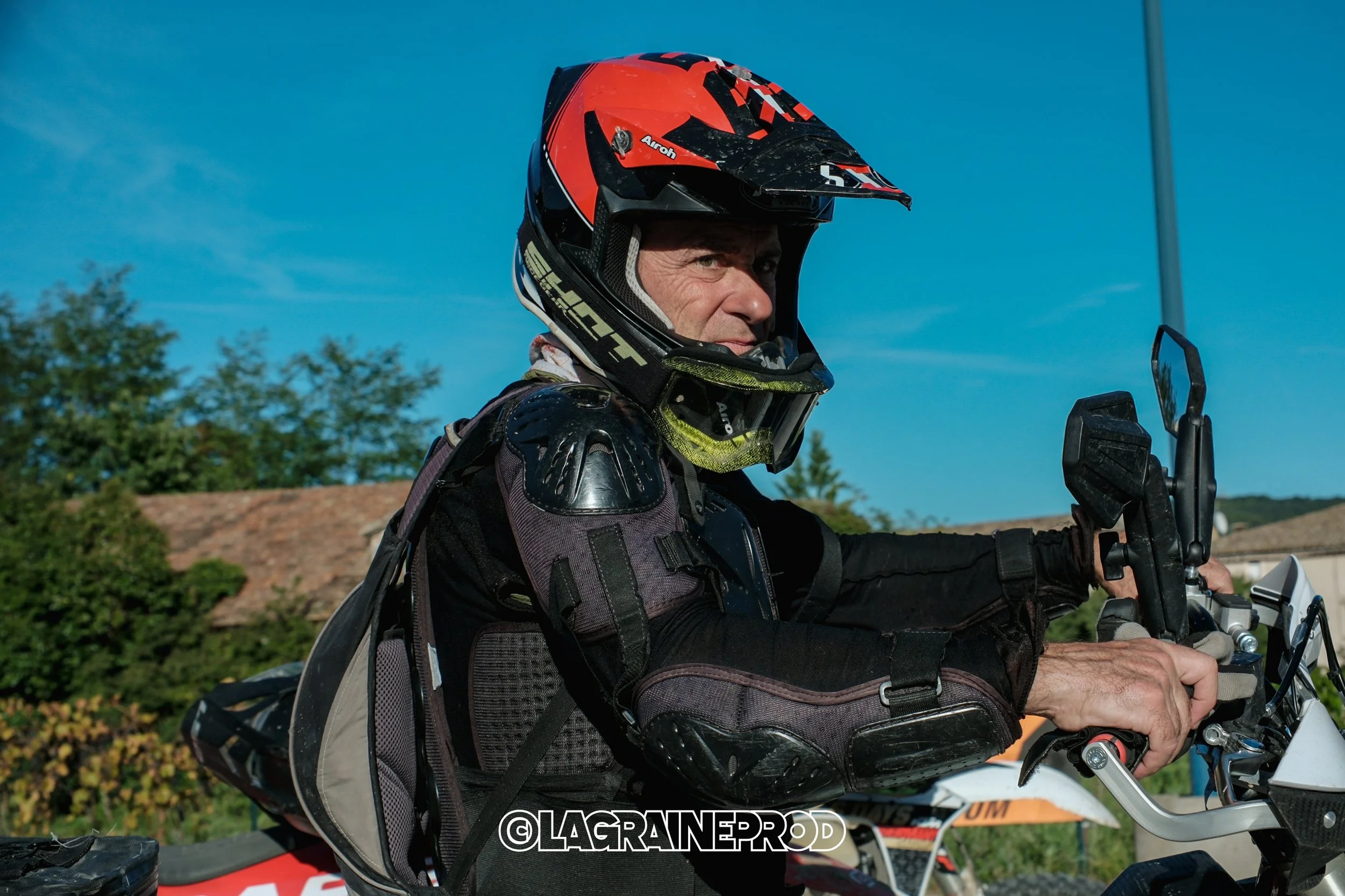 Un motocycliste en équipement de protection, portant un casque rouge et noir, au guidon d'une moto tout-terrain, en plein air sous un ciel bleu.