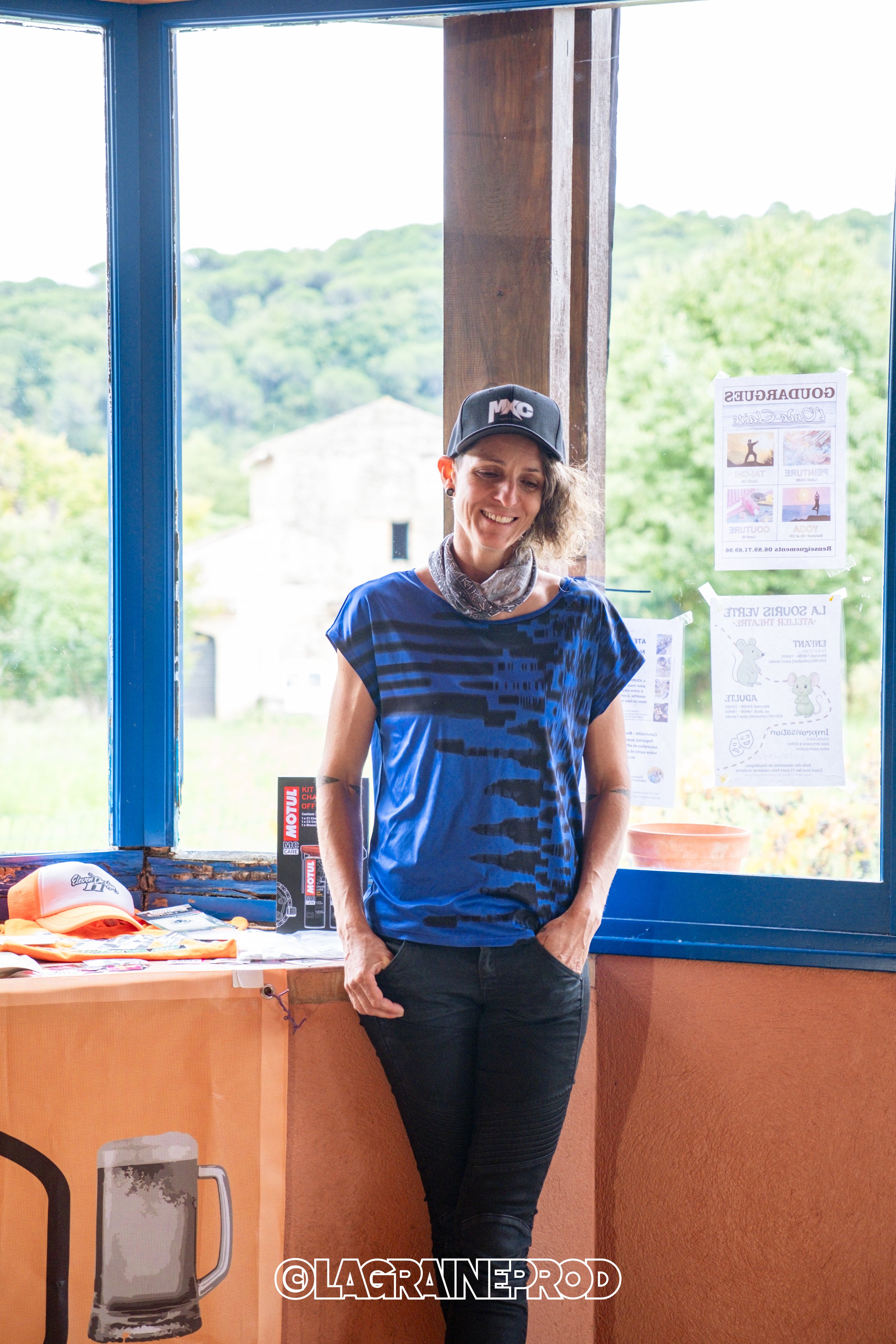 Une femme souriante portant un t-shirt bleu et une casquette noire, debout à l'intérieur près d'une fenêtre, avec des affiches et des objets promotionnels sur un comptoir à côté d'elle.
