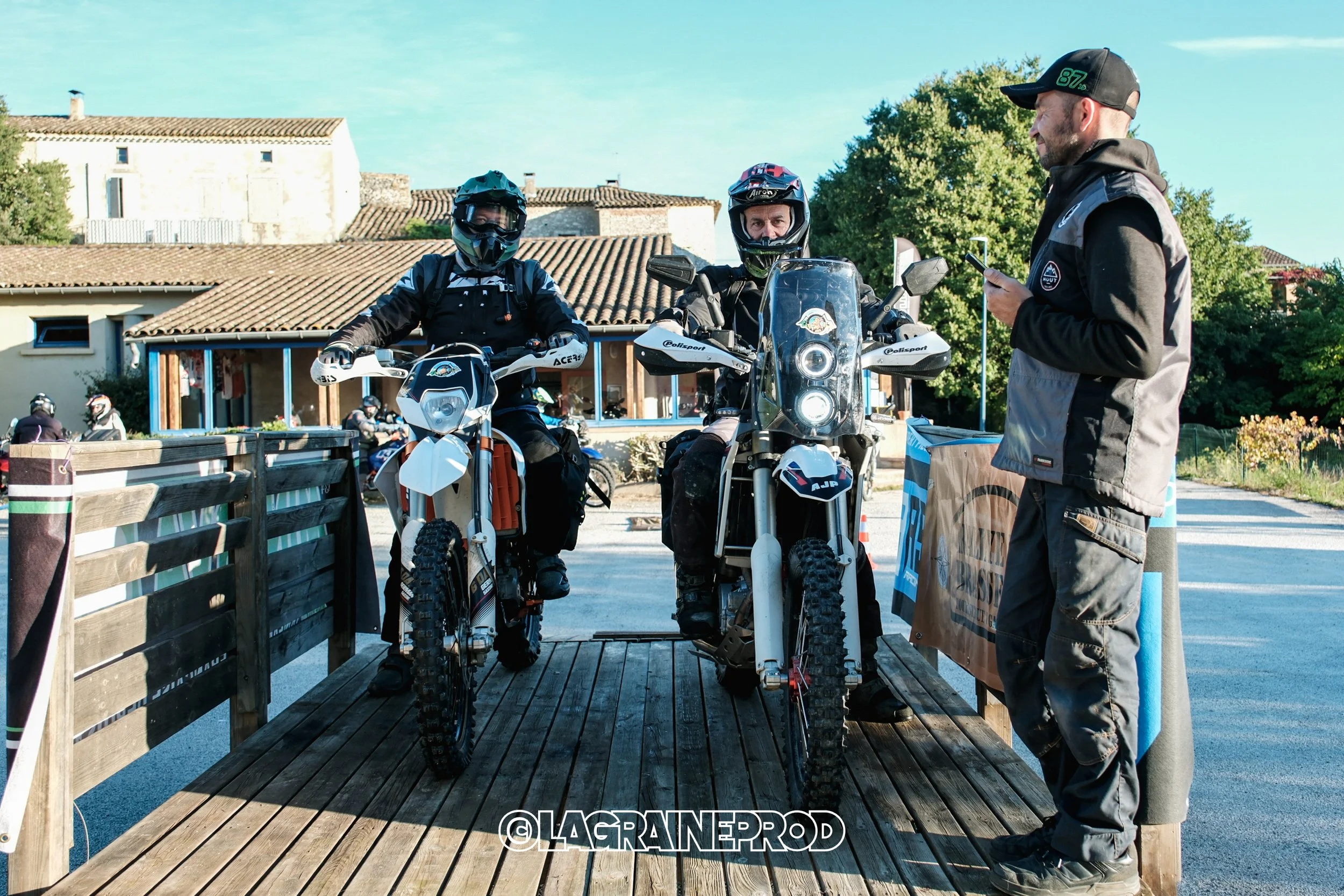 Deux motards en tenue de protection sur des motos tout-terrain, rencontrant un homme avec une veste à capuche, dans un environnement extérieur avec des bâtiments résidentiels et des arbres.