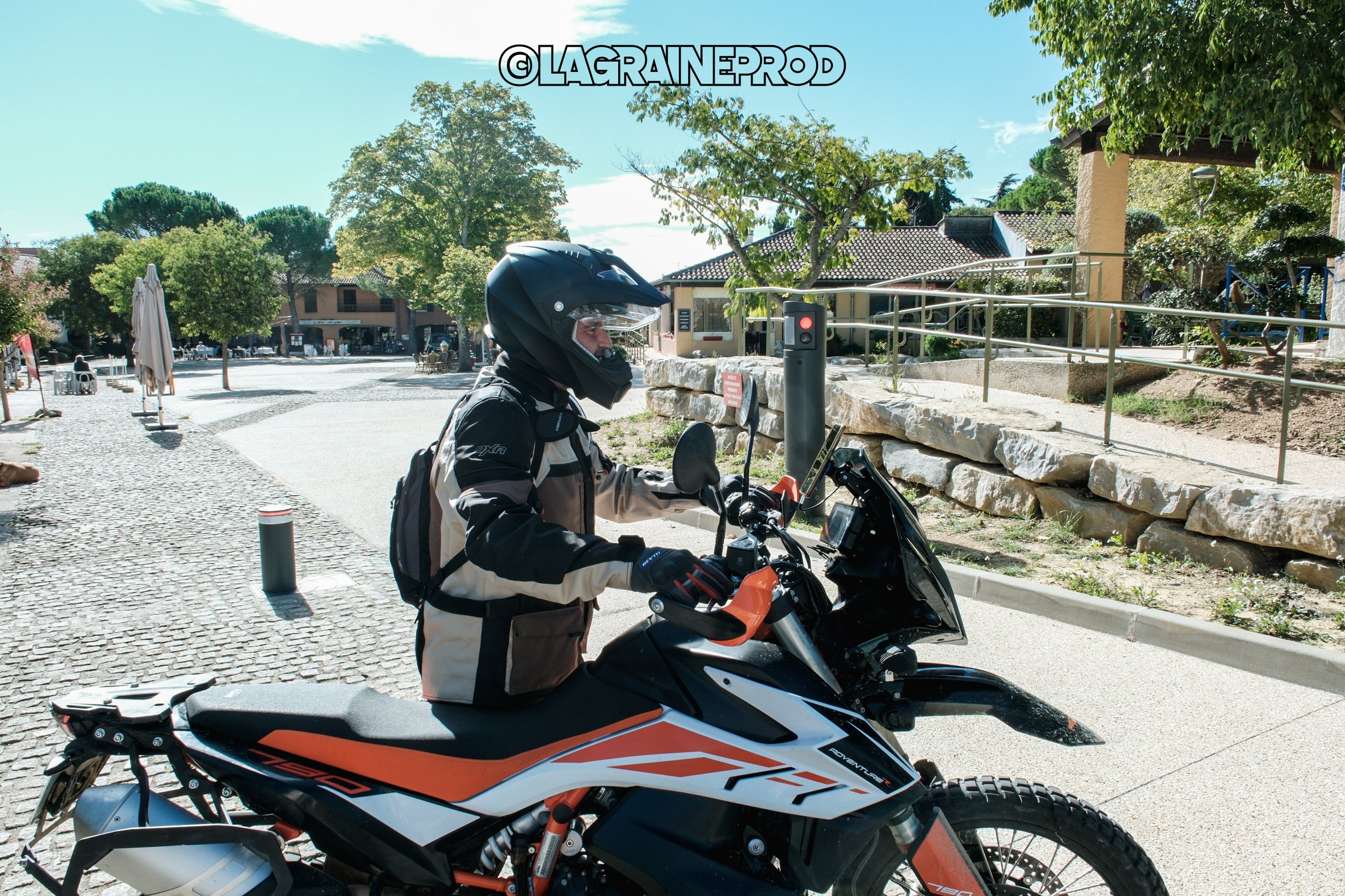 Un motard portant un casque et une veste, assis sur une moto tout-terrain orange et noir, dans une place piétonne en plein air sous un ciel ensoleillé.