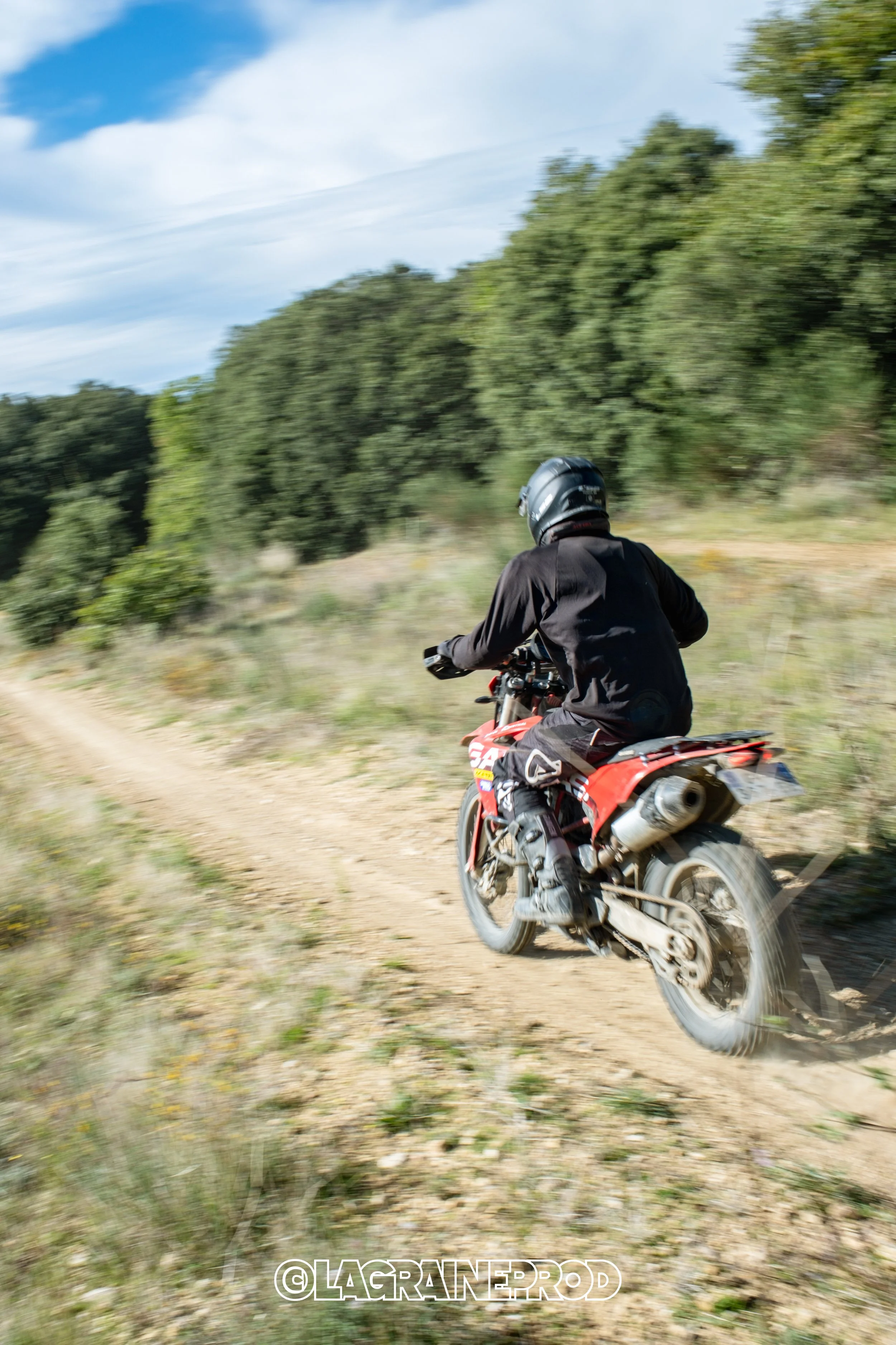 Un pilote en casque noir et veste noire roulant en moto sur un chemin de terre à travers une campagne verdoyante avec des arbres au loin et un ciel partiellement nuageux.