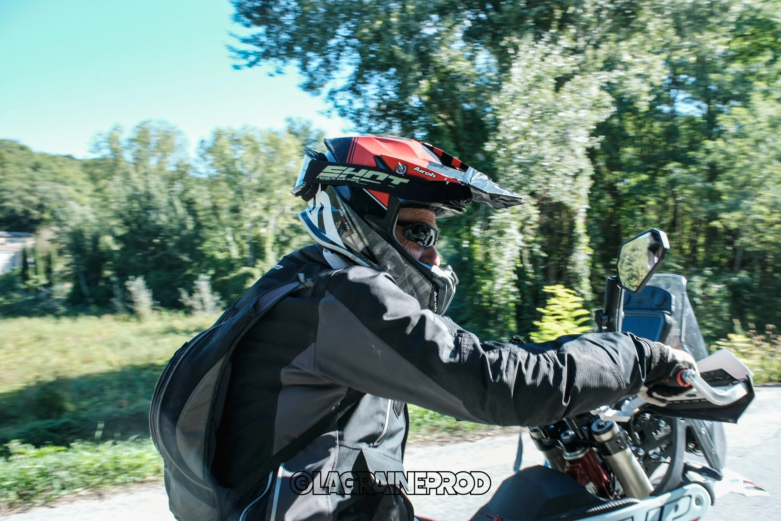 Un homme en casque de moto, lunettes et tenue de protection, roulant en moto dans la nature avec des arbres en arrière-plan.