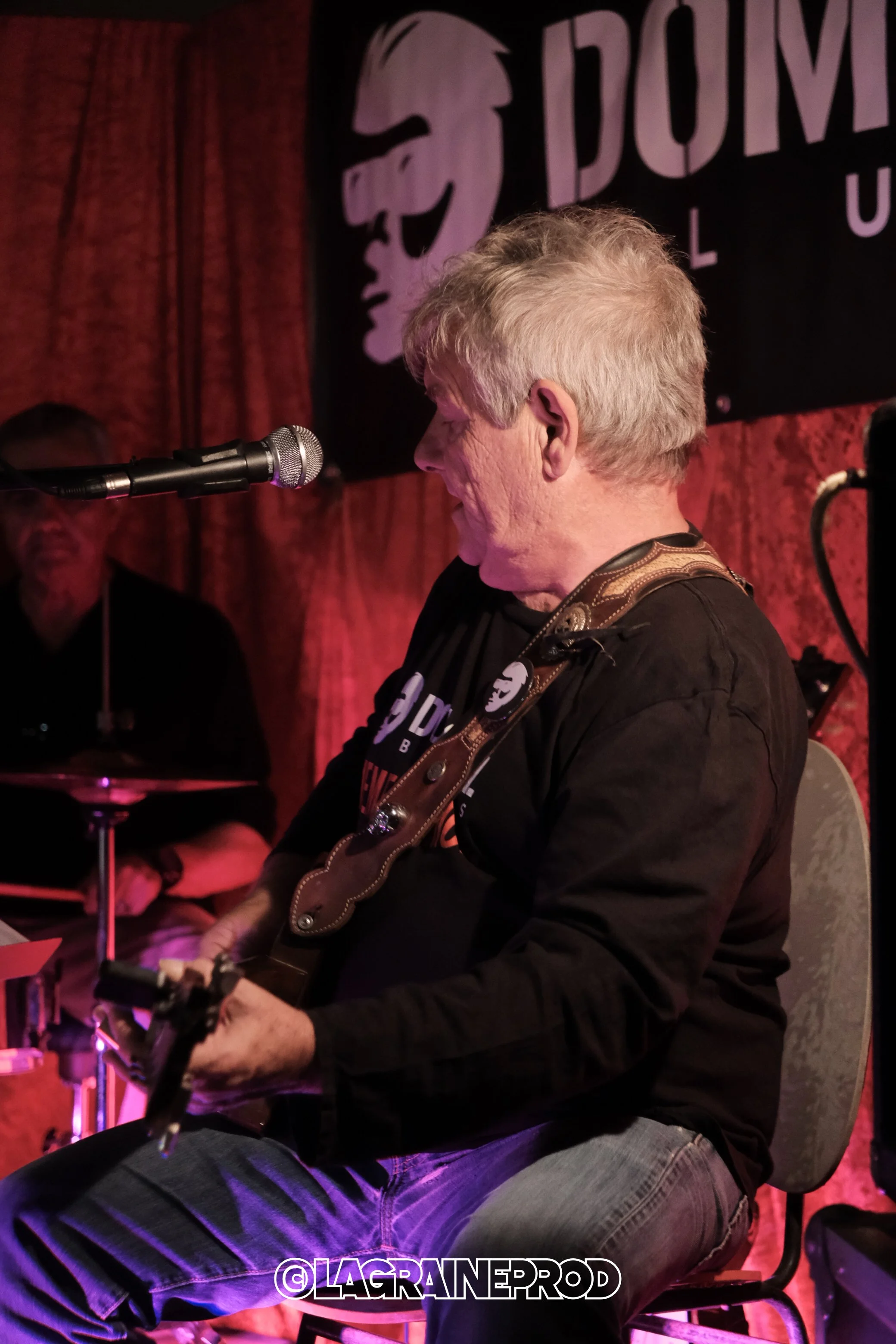 Un homme âgé joue de la guitare lors d'un concert dans une salle sombre avec un fond rouge, accompagné d'un batteur en arrière-plan, sur scène. 
