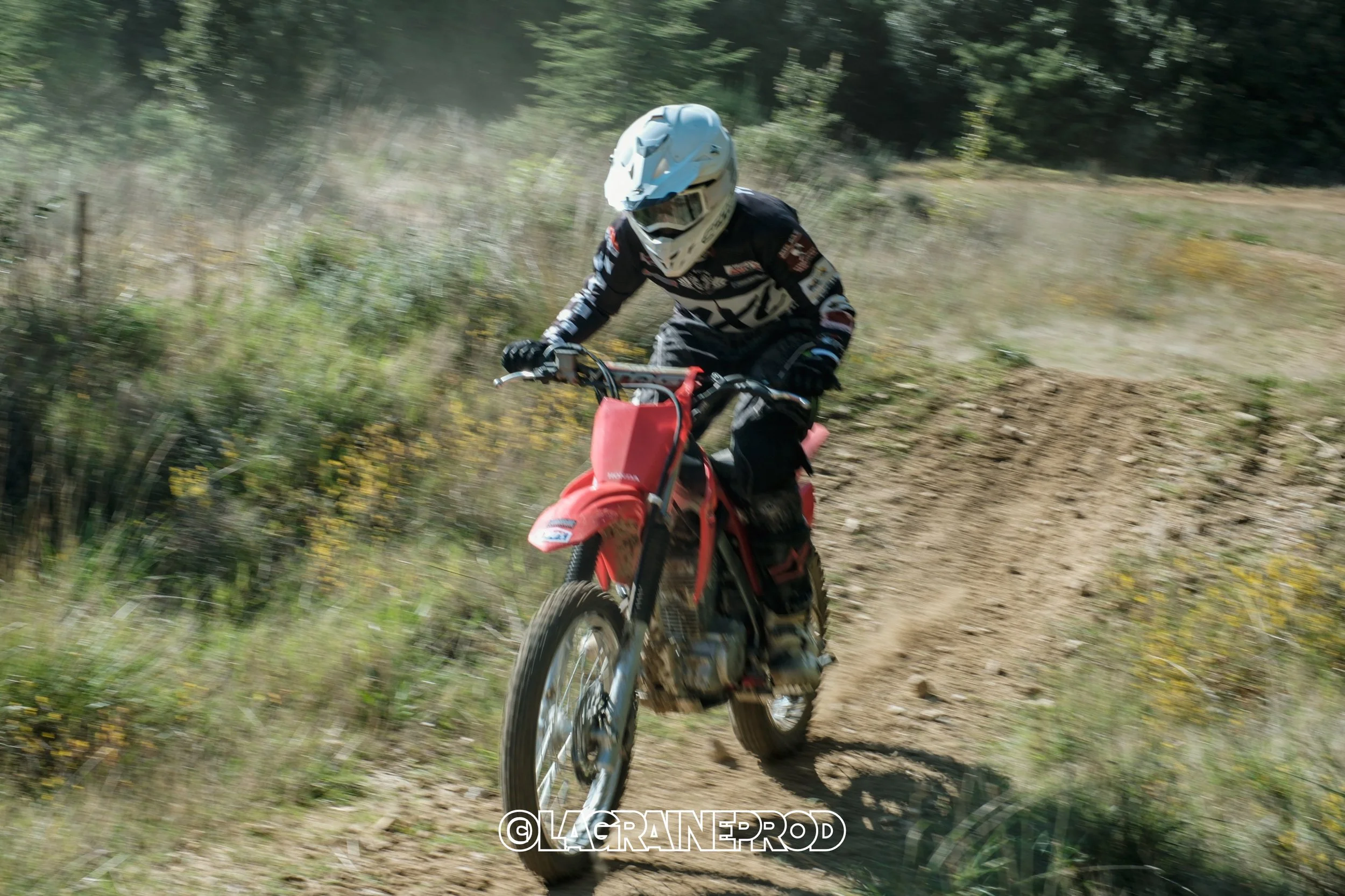Un motocycliste portant un casque blanc et une combinaison de motocross, roulant sur un sentier de terre en pleine nature.