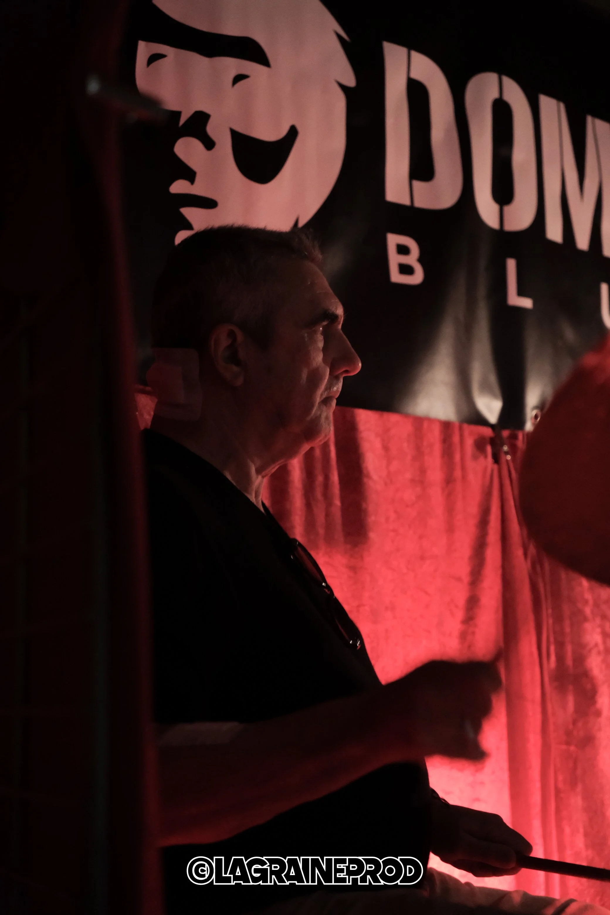 Un homme assis dans une scène sombre avec un fond noir et une bannière en arrière-plan affichant un visage de style caricature et le texte 'DOME BLU'. La scène est éclairée par une lumière rouge. L'homme porte un t-shirt noir et tient une baguette ou