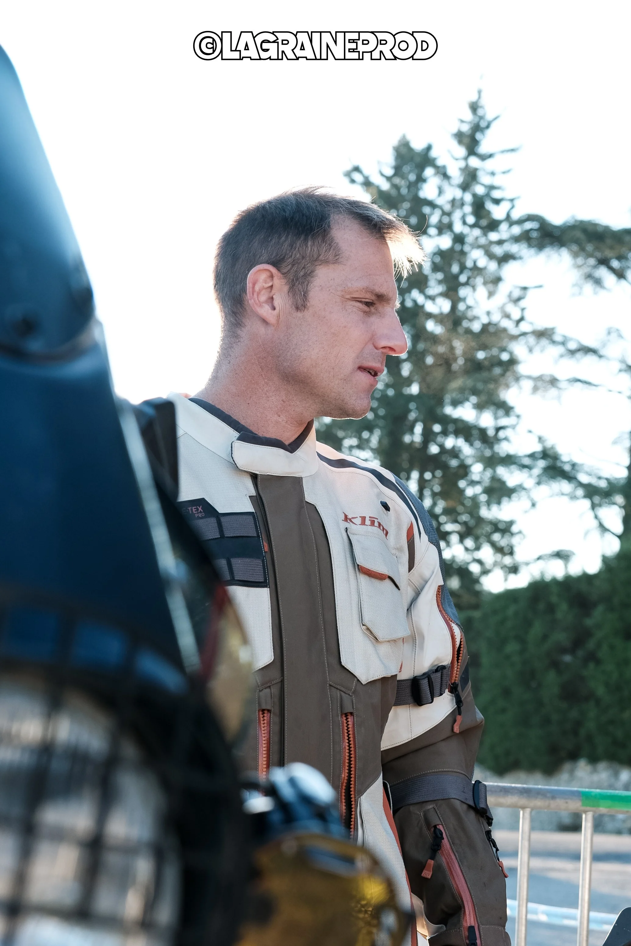 Un homme en tenue de motard regarde vers le bas, avec des arbres en arrière-plan et un casque de moto en avant-plan à gauche.