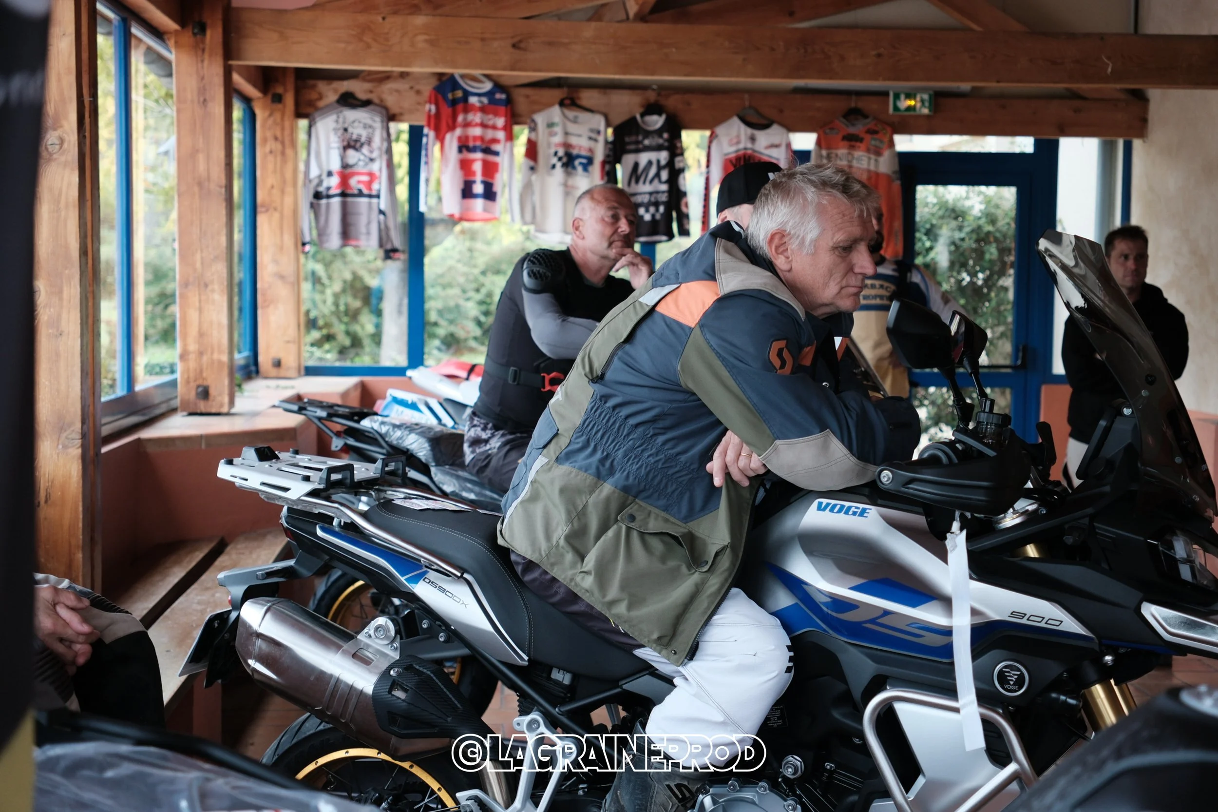 Homme assis sur une moto de sport à l'intérieur d'un boutique ou un espace d'exposition, avec d'autres personnes en arrière-plan, des affiches de vêtements de sport suspendues au plafond.