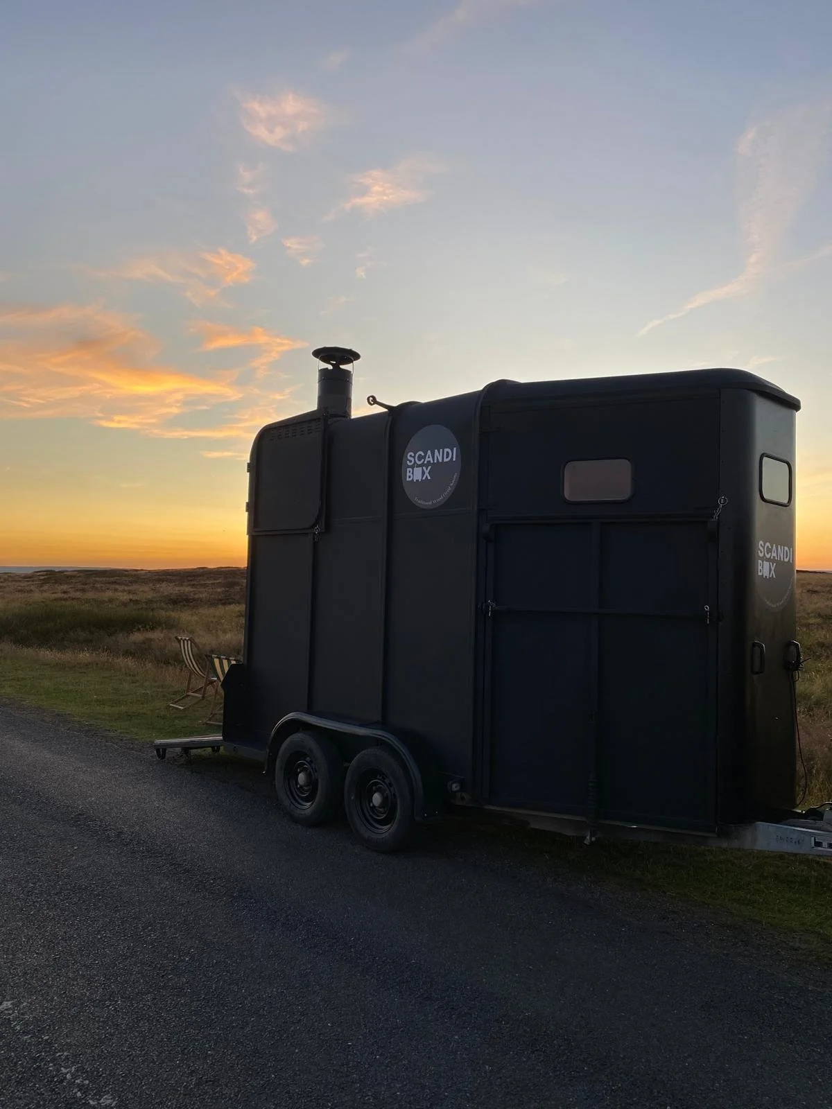 A black mobile tiny house trailer parked on the side of a rural road at sunset with a sky filled with orange and pink clouds.