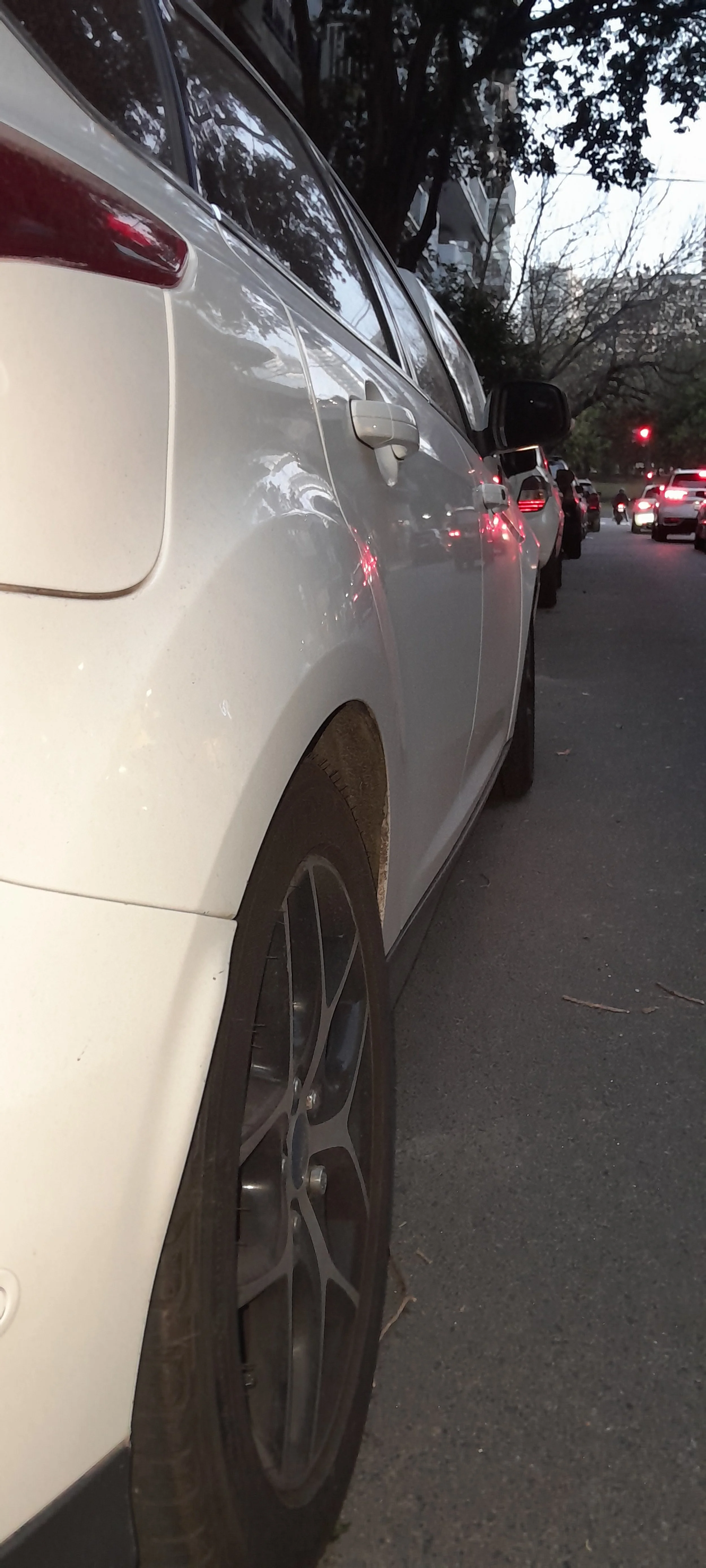Vista lateral de un coche blanco estacionado en la calle durante la tarde, con otros vehículos y árboles a su alrededor.