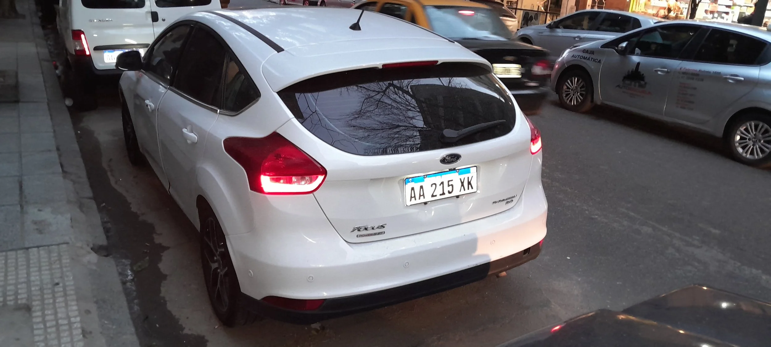 Vehículo blanco modelo Ford Focus estacionado en la calle con otros carros en movimiento al fondo.
