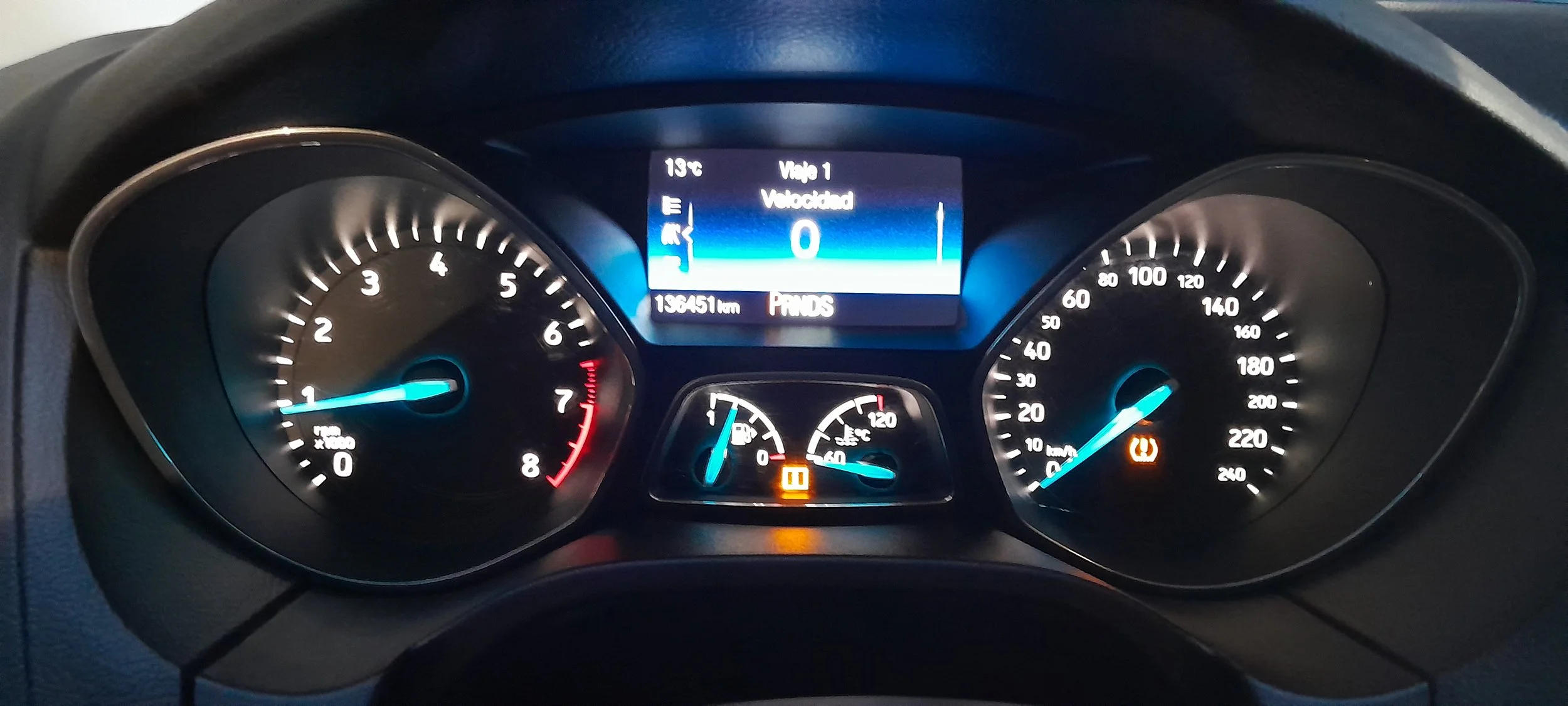 Tablero de instrumentos de un automóvil que muestra una velocidad de 20 km/h, una temperatura de 13°C, una lectura de odómetro de 139,451 km, y luces de advertencia encendidas, incluyendo el freno de mano y la rueda pinchada