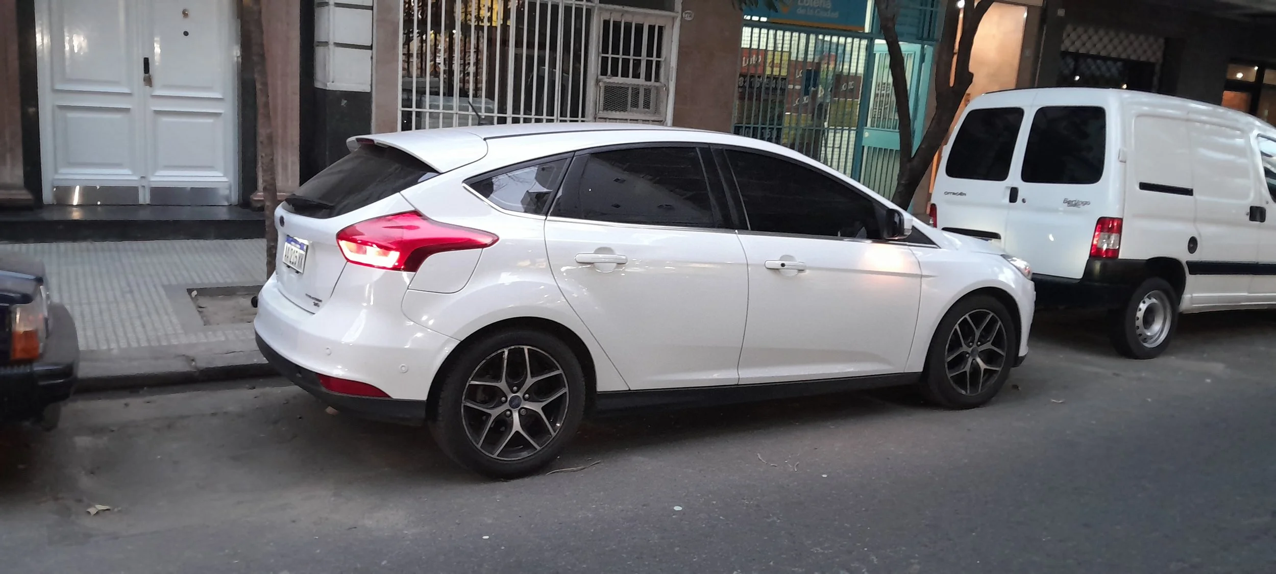 Auto sedán blanco estacionado en la calle, junto a una camioneta blanca, con edificios y rejas en el fondo.