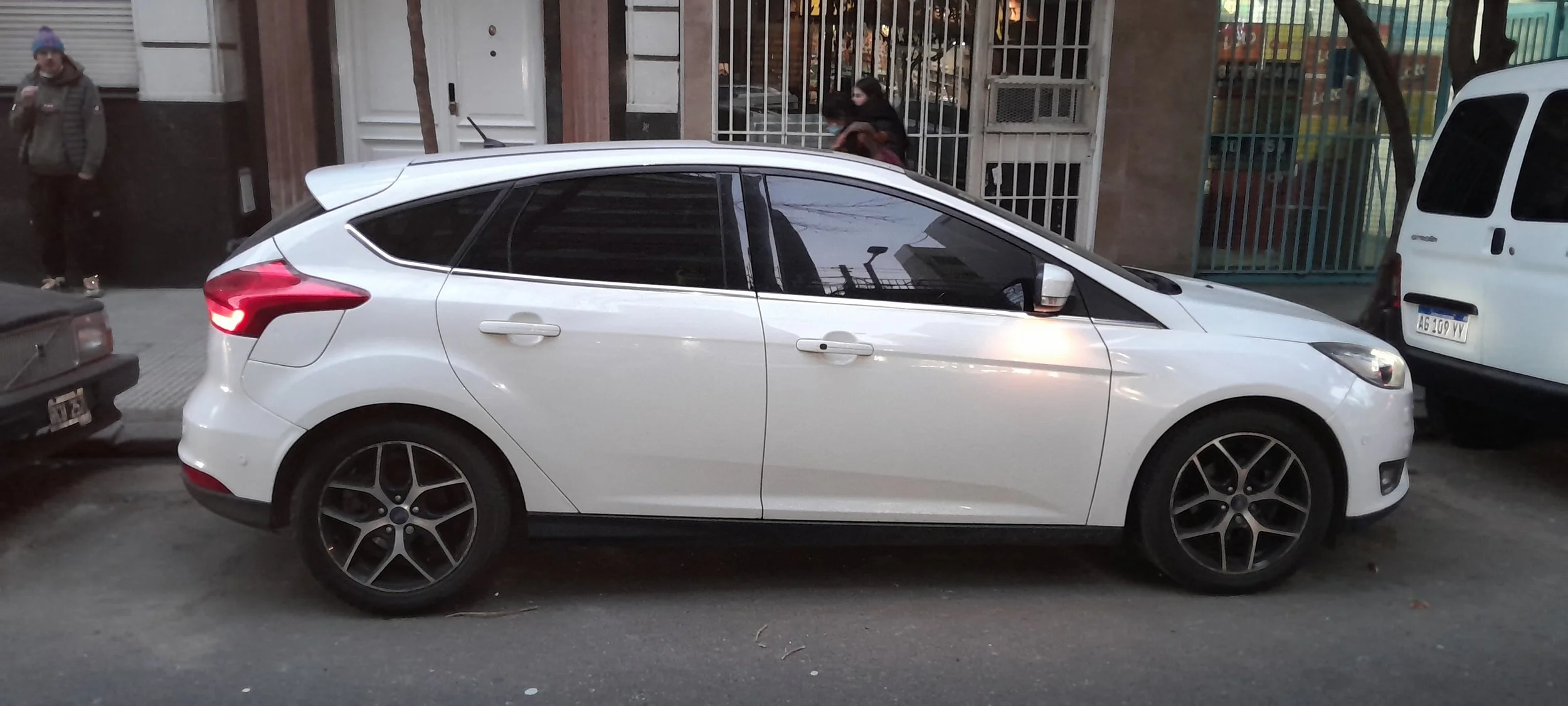 Auto compacto blanco estacionado en la calle, con edificios y otras personas y vehículos en el fondo.